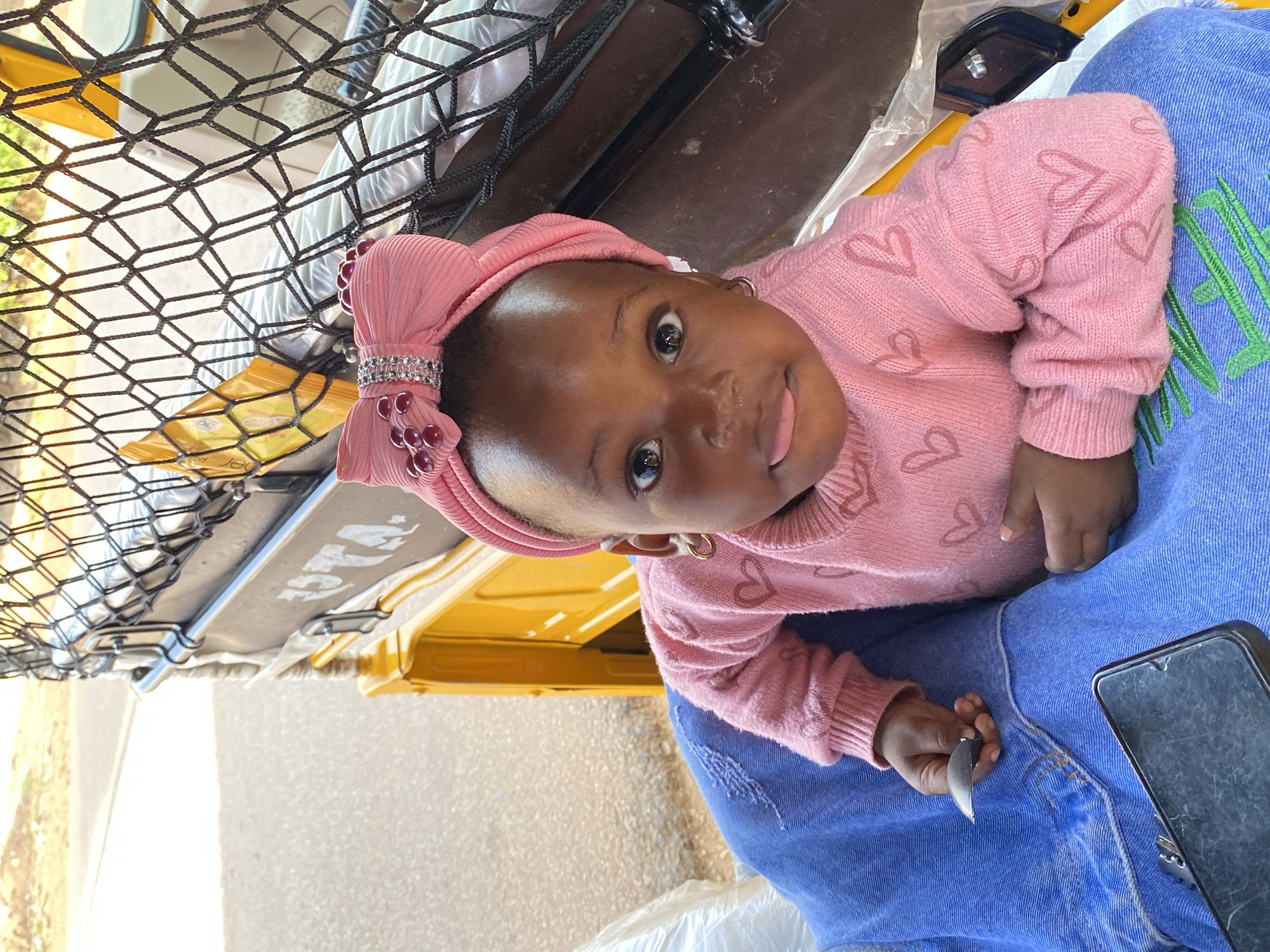 The image, taken in Vom, Nigeria, presents a close-up, portrait-oriented view of a baby held on an adult's lap, likely within or next to a vehicle. The baby, with dark skin, looks directly into the camera with wide, curious eyes and a slightly open mouth, revealing a glimpse of her tongue. She is adorned with a textured pink headwrap featuring a large bow decorated with rhinestones and small red/pink beaded accents, and wears small gold hoop earrings. Her attire also includes a cozy pink sweater patterned with numerous small hearts.
The adult's lap, wearing blue, possibly corduroy, jeans with some distressed areas, supports the baby. A black smartphone in a protective case is visible resting on the adult's lap to the baby's right. The baby's left hand grasps a small, shiny dark metal object, perhaps a key or a simple tool.
The background reveals elements of an outdoor setting during daytime, with bright, likely sunny, ambient light. A black net with a diamond pattern is prominent in the upper left. Behind the net and partially to the baby's right, a bright yellow structure, characteristic of a local public transport tricycle (often called 'Keke NAPEP' in Nigeria), is visible. On a black panel behind the net, the letters "P.S.T.A." are clearly legible, suggesting a transport service or association. The scene captures a candid moment, highlighting the baby's engagement and curiosity.