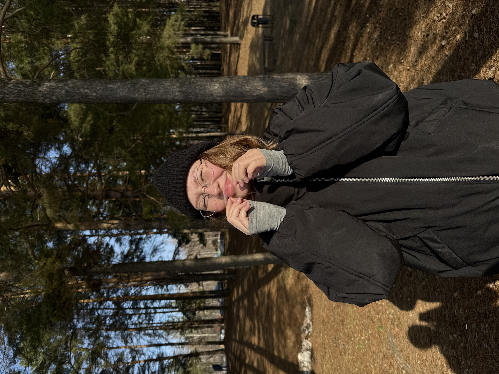 A person wearing a black beanie, round glasses, and a black jacket is standing outdoors in a wooded area. The person has their hands near their face, possibly touching their lips. The background consists of tall pine trees and fallen leaves on the ground. In the distance, there is a structure that resembles a bench or trash can. The scene is illuminated by natural daylight. This picture was most likely taken in the city of Chaikovsky, Russia.