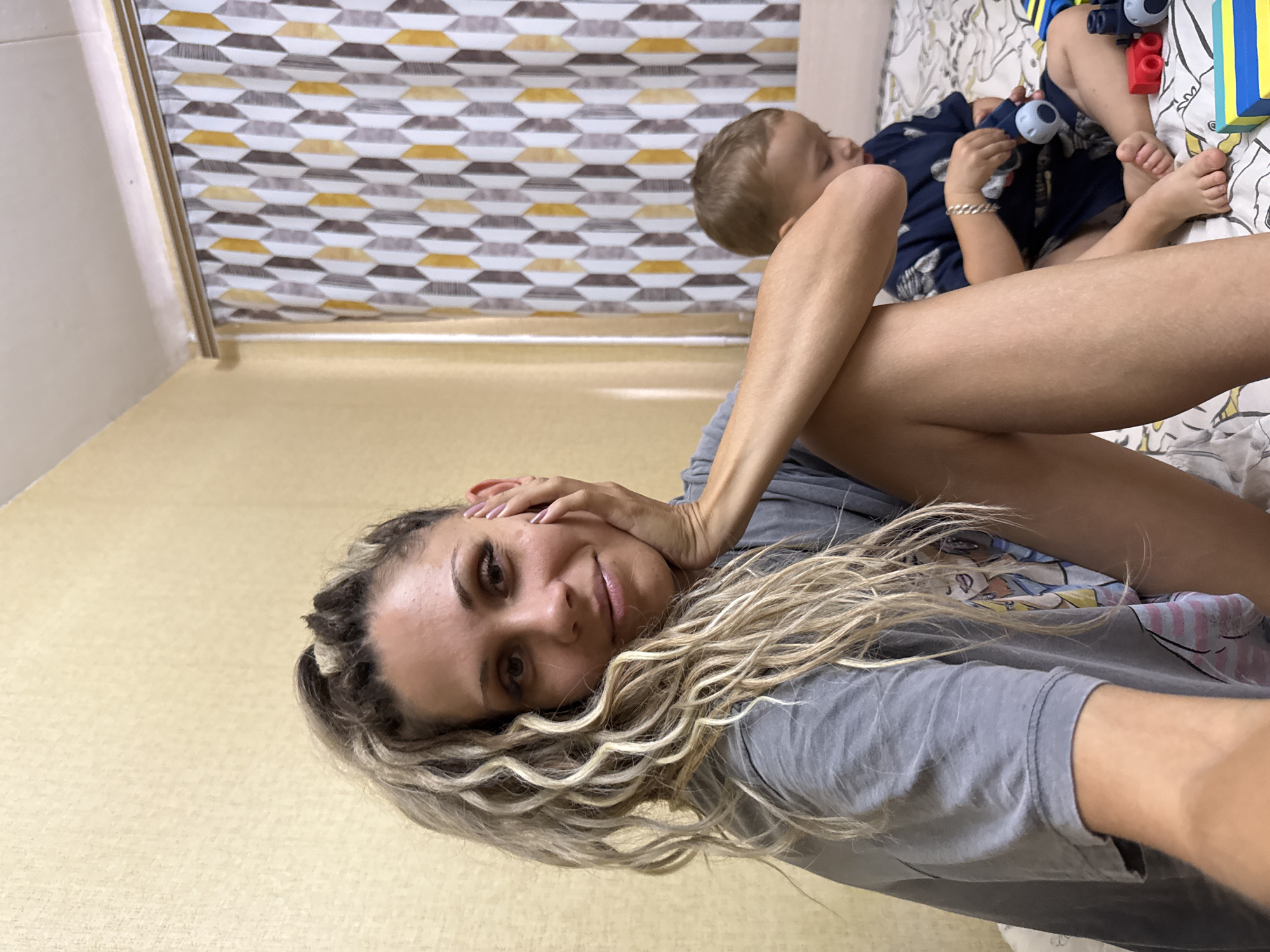 In Rusanovo, Russia, a woman with blonde, curly dreadlocks and a grey t-shirt is sitting on a bed, her hand resting on her cheek. She is looking towards the camera with a slight smile. Next to her, a young boy in a dark blue shirt is playing with toys on the bed. The room has yellow-patterned wallpaper and a curtain with a geometric design.