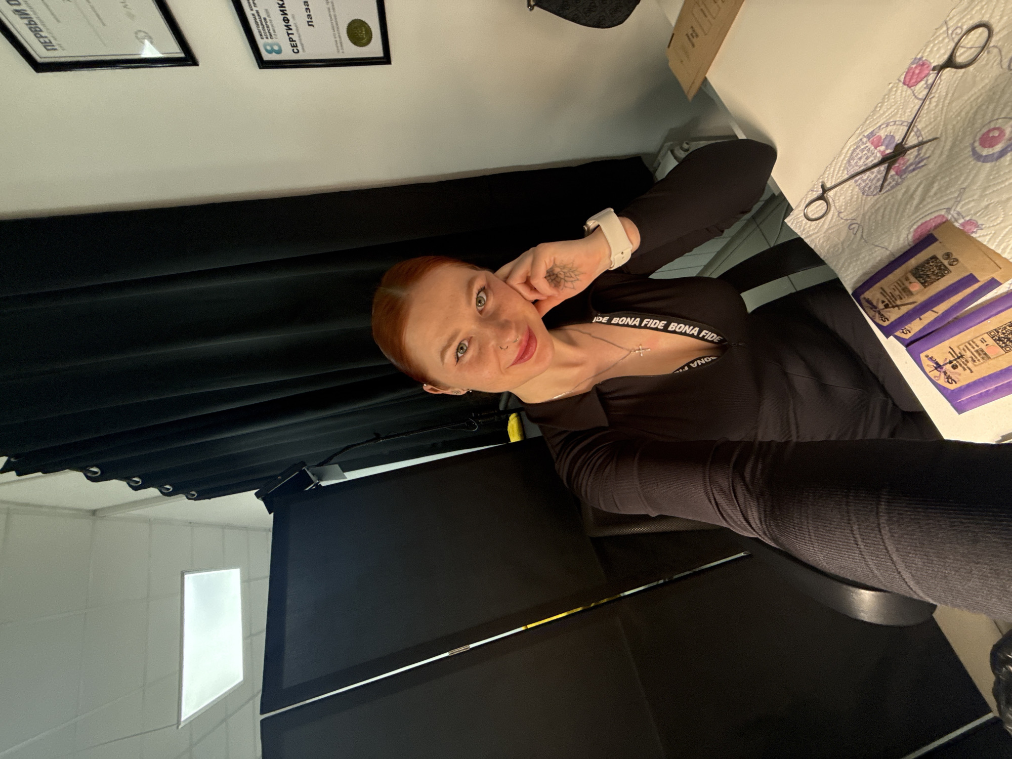 A red-haired female with freckles, a nose piercing, a lotus tattoo on her left hand, and wearing a dark, long-sleeved top with a visible "BONA FIDE" strap and a cross necklace, is pictured in a selfie. She is seated at a white desk, smiling, with her left hand resting on her cheek, in what appears to be a professional studio located in Rostov-on-Don, Russia. On the white desk, a patterned paper towel displays a pair of piercing forceps and three individually packaged sterile items, labeled "Steel Power," next to an unfolded brown paper envelope. The background features dark privacy curtains, a white drop ceiling with a rectangular fluorescent light, and a white wall displaying two framed certificates in Russian. One partially visible certificate is titled "ПЕРВЫЙ О" (First O) and another is labeled "СЕРТИФИКАТ" (Certificate), with text indicating "Лаза" (Laser) and "ПИРСЕРСКАЯ" (Piercing). A black office chair and a black bag are also visible in the immediate environment.