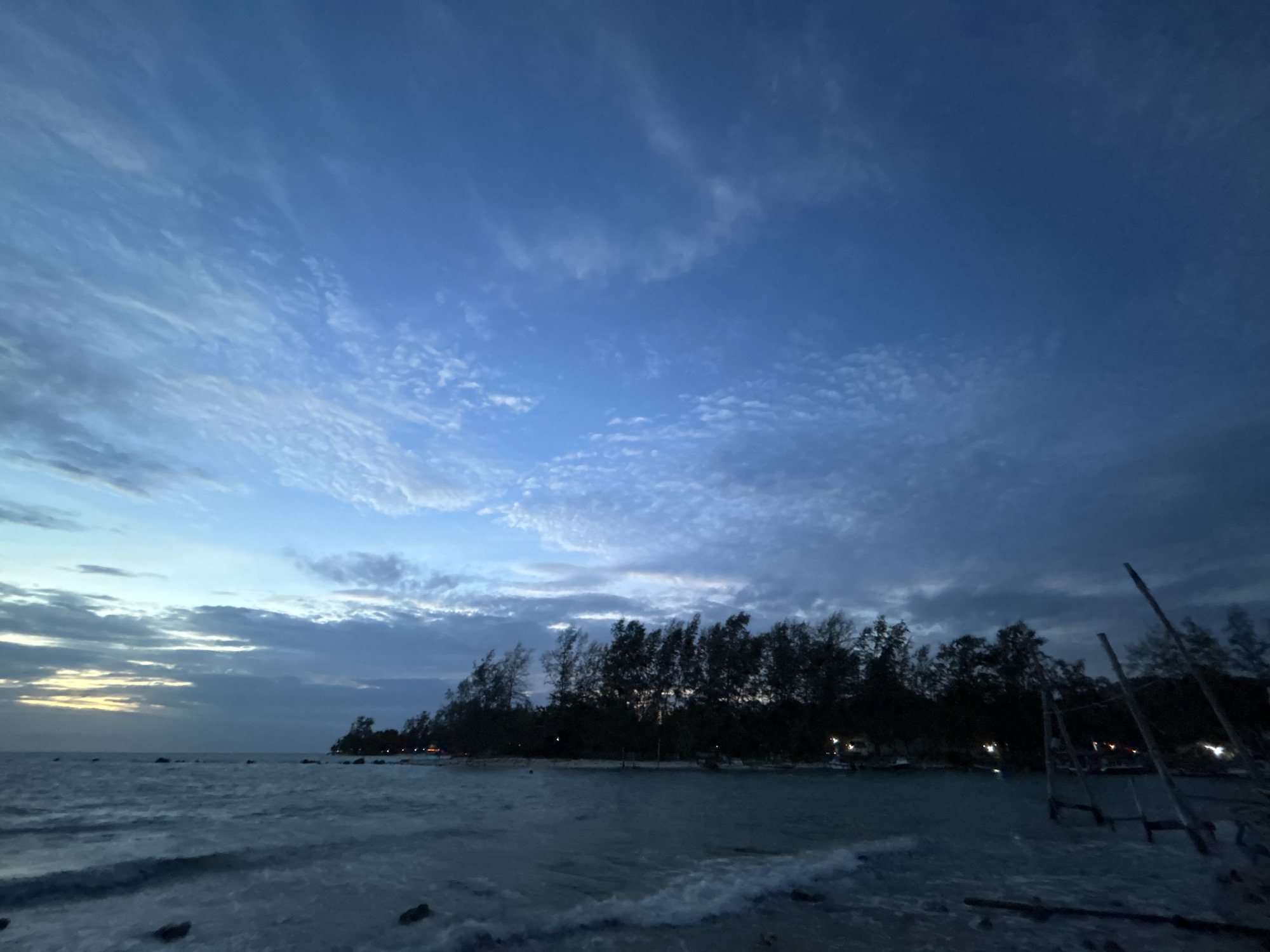 A serene twilight scene unfolds on a beach in the Ko Pha-ngan District of Thailand. The sky, a gradient of deep blues and soft grays, is streaked with wispy clouds, hinting at the remnants of daylight. In the foreground, gentle waves lap against the shore, their surface reflecting the subdued light. The water is a tranquil turquoise, transitioning to a darker hue further out.
Along the shoreline, a dense line of trees stands silhouetted against the sky, their branches swaying slightly in the evening breeze. Beyond the trees, small lights flicker, suggesting human habitation or activity, perhaps a resort or local dwellings. To the right of the frame, a rustic wooden structure, possibly a jetty or a boat mooring, juts out into the water, its wooden posts stark against the dimming sky. The overall atmosphere is one of peace and quiet, characteristic of a tropical beach at dusk.