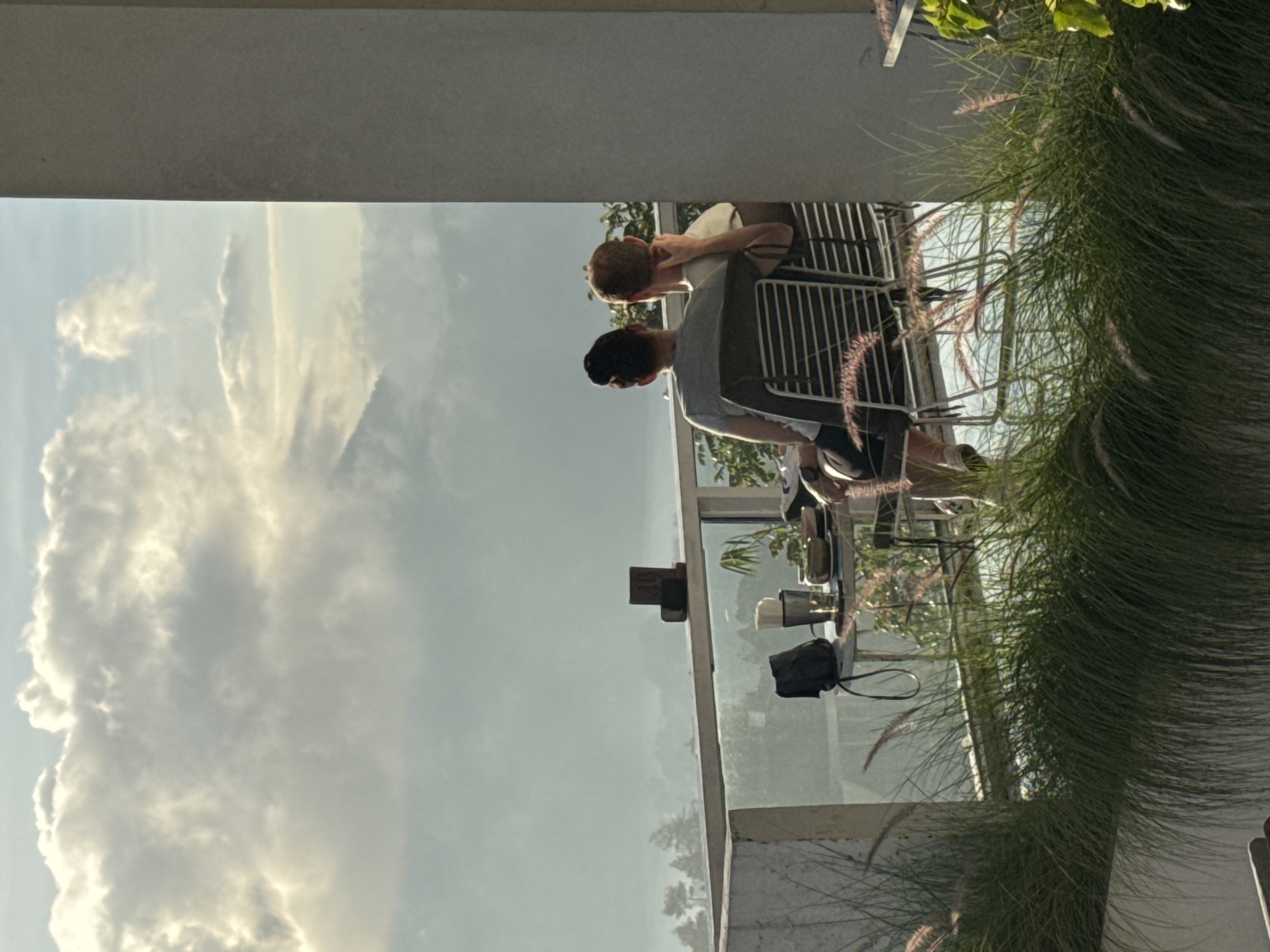 Scene: A cafe in Kedisan, Indonesia, viewed through a glass-enclosed balcony.
People: Two individuals are present. One is a young boy with reddish hair, leaning towards another person, presumably engaged in conversation. The other person, possibly an adult, is wearing a grey shirt.
Objects: Chairs and tables are arranged on the balcony. A purse, cup, and possibly other items are on the table.
Environment: The scene is outdoors, with a view of cloudy sky. Pampas grass is near the right edge of the balcony.