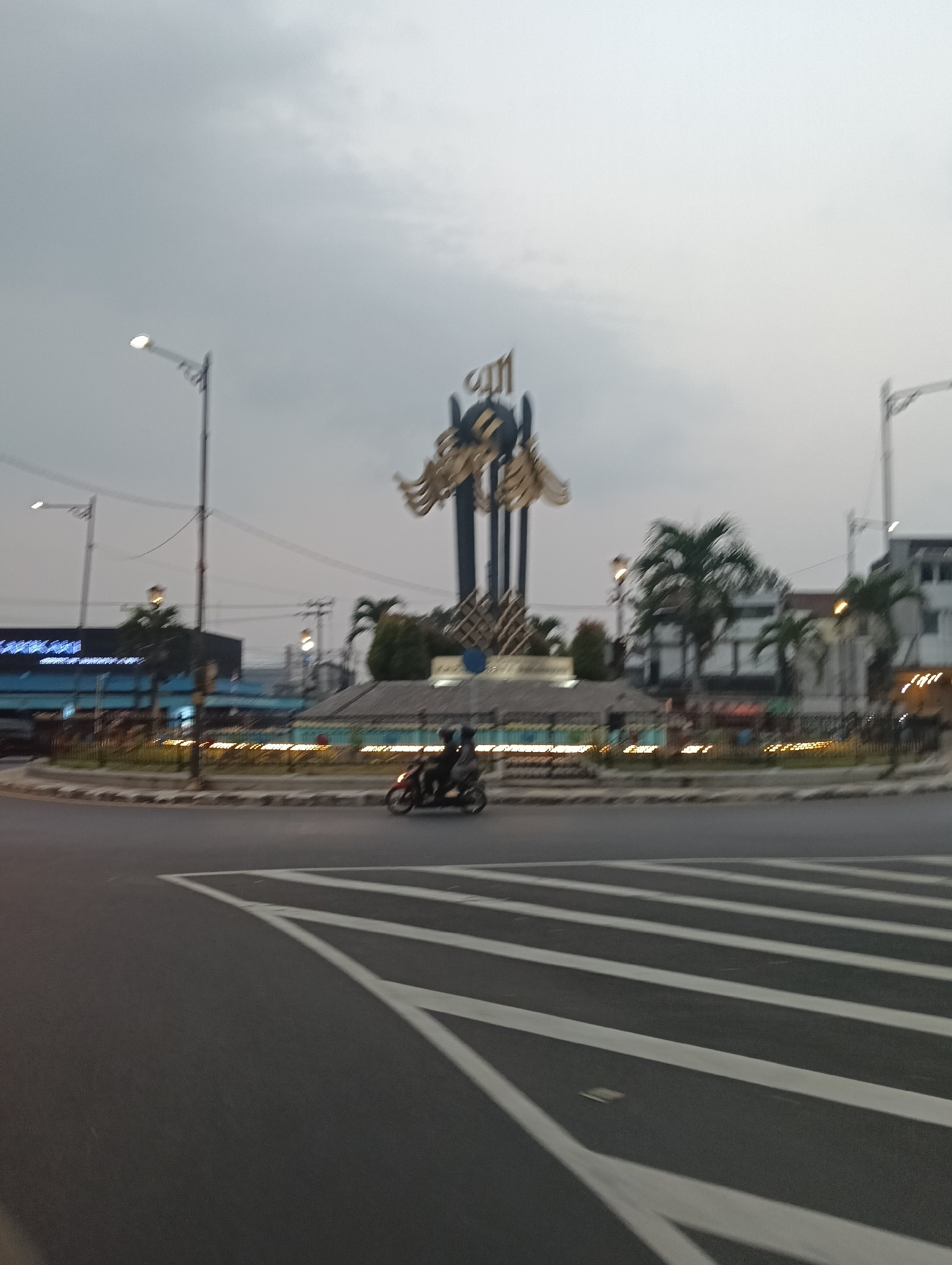 The image shows a street scene in Tasikmalaya, Indonesia, likely at dusk or early morning due to the overcast sky and streetlights being illuminated. In the center of the image is a large, elaborate monument with Arabic calligraphy at the top, possibly indicating a religious or historical significance. The monument is surrounded by palm trees and some low-lying vegetation. A road curves around the monument, with white lane markings visible on the asphalt. A motorcycle with two riders is traveling on the road in the middle ground. In the background, there are buildings, including what appears to be a commercial establishment with illuminated signage, and streetlights. The overall impression is of an urban environment at the beginning or end of the day.