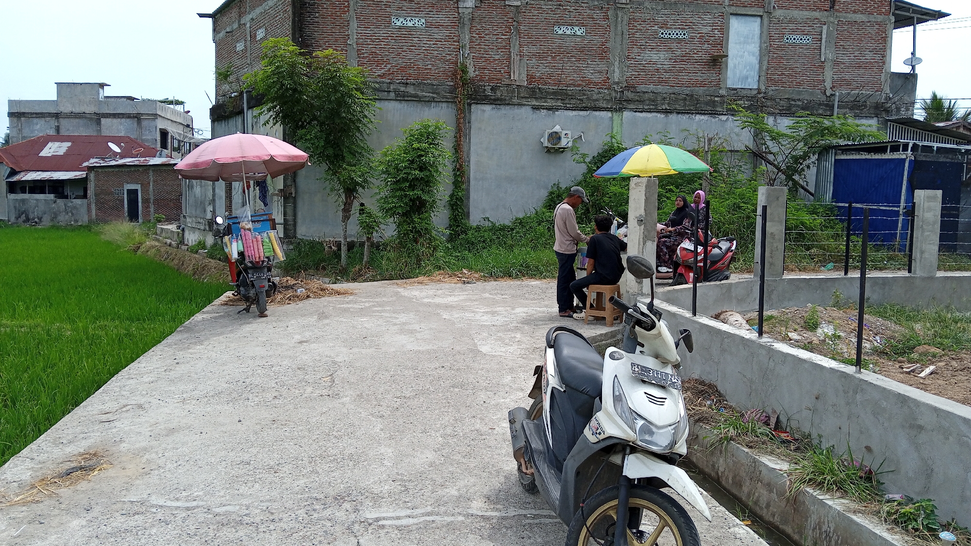In Geulangganggampong, Indonesia, a concrete path runs alongside a vibrant green rice paddy. A parked motorcycle with an attached cart laden with colorful items, possibly ice cream or drinks, stands under a pink umbrella. Further along the path, two men are engaged in an interaction, one standing and the other seated on a stool, beneath a yellow and blue umbrella. Another motorcycle is parked nearby, and two women are seated on it. A partially constructed concrete barrier runs along the right side of the path, with sparse vegetation growing in and around it. In the background, there are buildings constructed of brick and concrete, with varying rooflines and architectural styles. The overall scene depicts a casual street-side commercial activity in a semi-urban or rural Indonesian setting.
