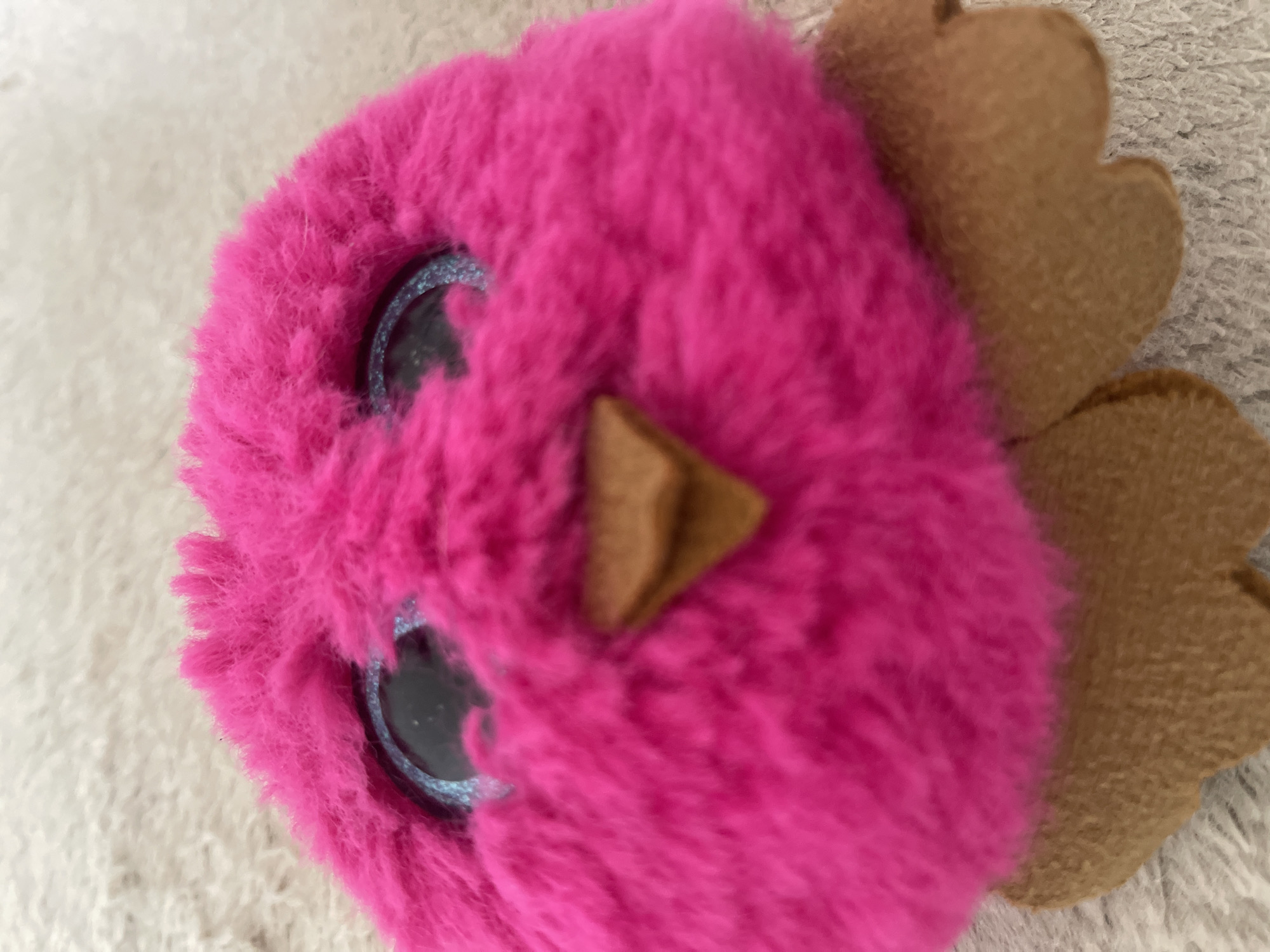 The image is a close-up, slightly out-of-focus shot of a fluffy, bright pink plush toy resembling a bird. The toy has large, sparkly blue eyes and a small, brown beak. It is positioned on a textured, light-colored surface, possibly a carpet or blanket. Two brown, wing-like appendages are visible at the bottom of the toy. The specific location in Depok, Indonesia, cannot be determined from the provided image.