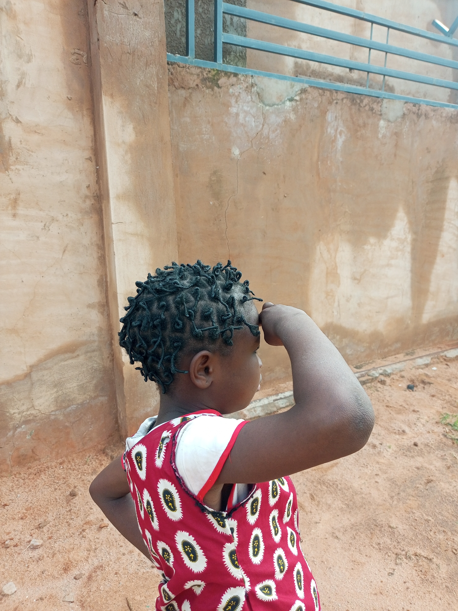 A medium shot captures a young person from the shoulders up, facing away from the camera and to the right, with their right arm raised to their forehead. The person has dark skin and their hair is styled in numerous small, dark twists. They are wearing a red dress with a white, black, and yellow oval pattern. The background consists of a textured, light brown wall and a metal railing above it. The ground in front of the wall is sandy and dry. This scene appears to be outdoors, possibly in a residential area in Tempure, Nigeria.