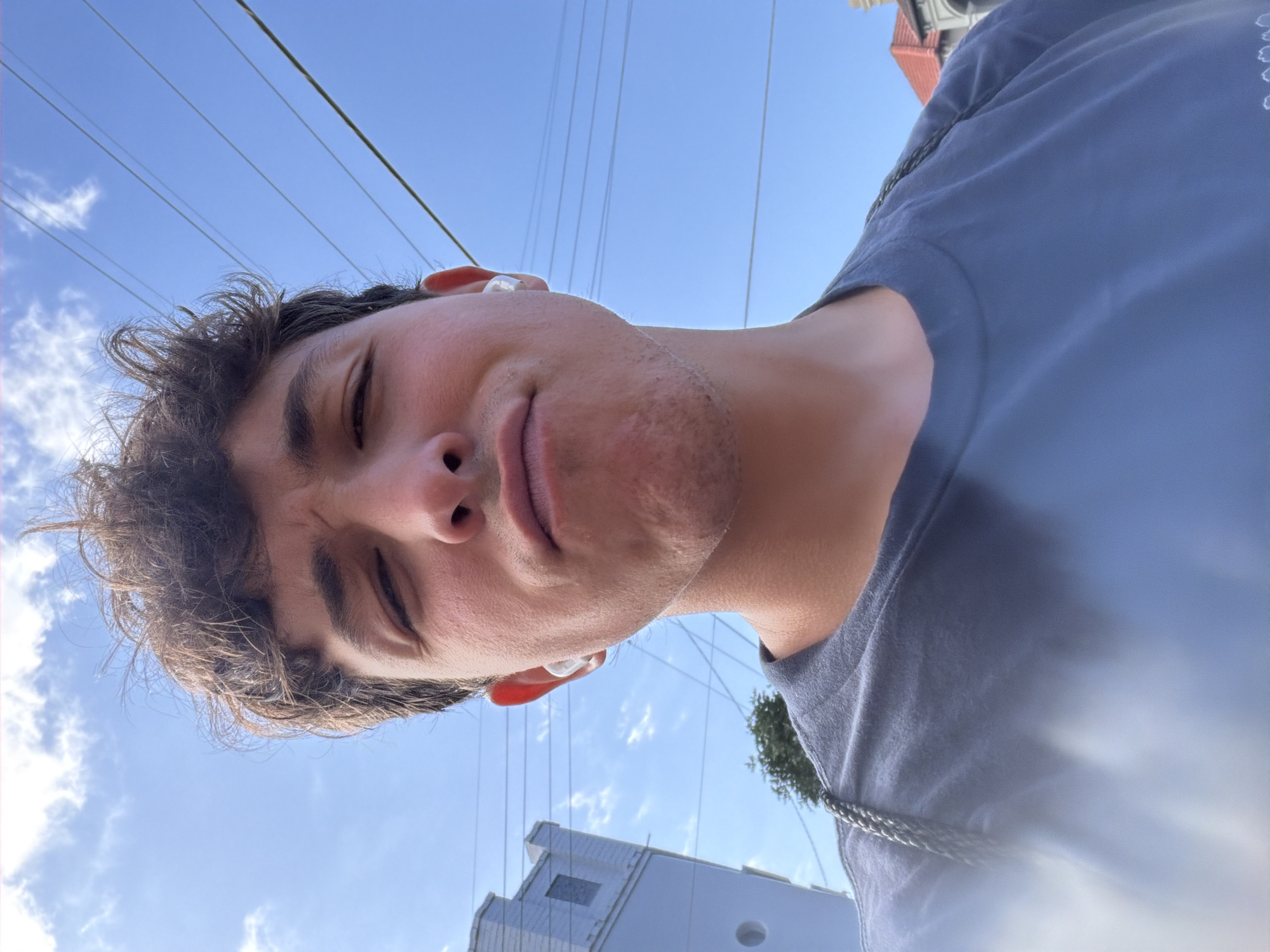A young man with dark, curly hair and a slight stubble is taking a selfie outdoors. He is wearing a blue t-shirt and white earbuds. His expression is a playful pout. In the background, the sky is blue with scattered clouds, and power lines are visible. A light-colored building and a tree are also present. The image appears to have been taken on a sunny day in Clovelly, Australia.