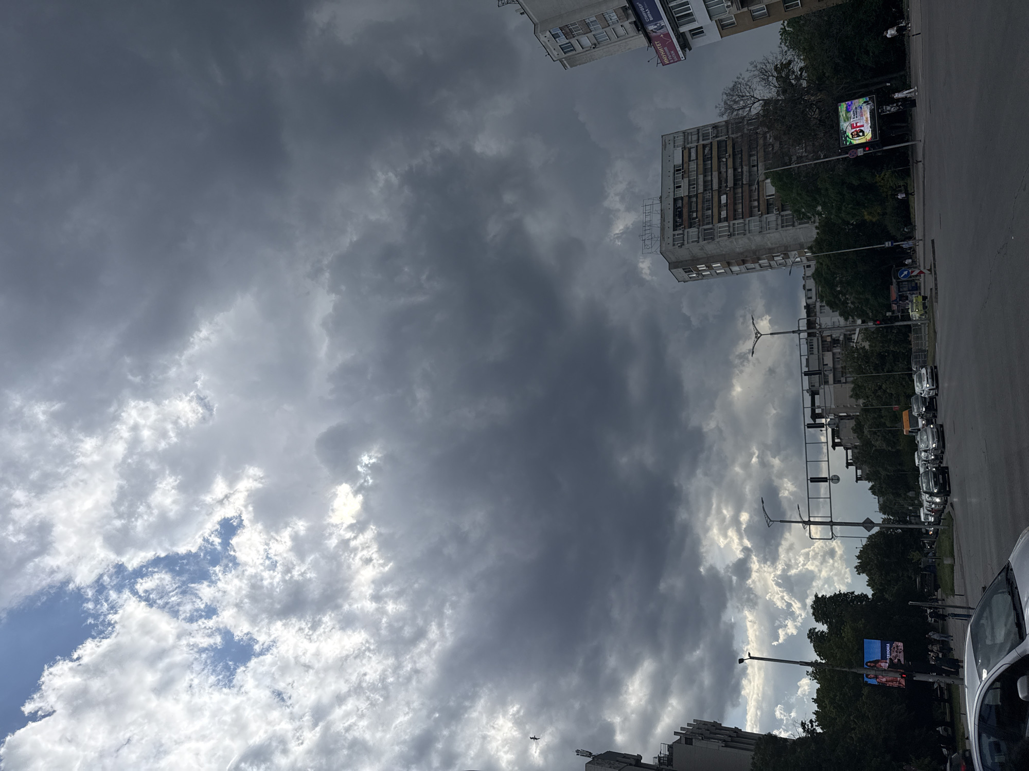 This is a street-level, eye-level, medium shot of a city intersection in Plovdiv, Bulgaria, under a dramatic sky with heavy, dark storm clouds and patches of sunlight breaking through. The foreground shows the asphalt of a road with a silver car in the lower left corner. In the middle ground, traffic is visible, including several cars and a yellow taxi, moving through the intersection. Traffic lights and street lamps are present. On the left side of the road, there are trees and a large digital billboard displaying an advertisement. Further back, on the right side of the intersection, stands a tall, multi-story apartment building with a weathered facade. Another, less tall building is visible behind it. In the far distance, more buildings and trees can be seen. The sky is a dominant feature, filled with large, textured clouds ranging from white and grey to dark charcoal, suggesting an impending or passing storm. A solitary bird is in flight against the cloudy sky on the left side of the frame.