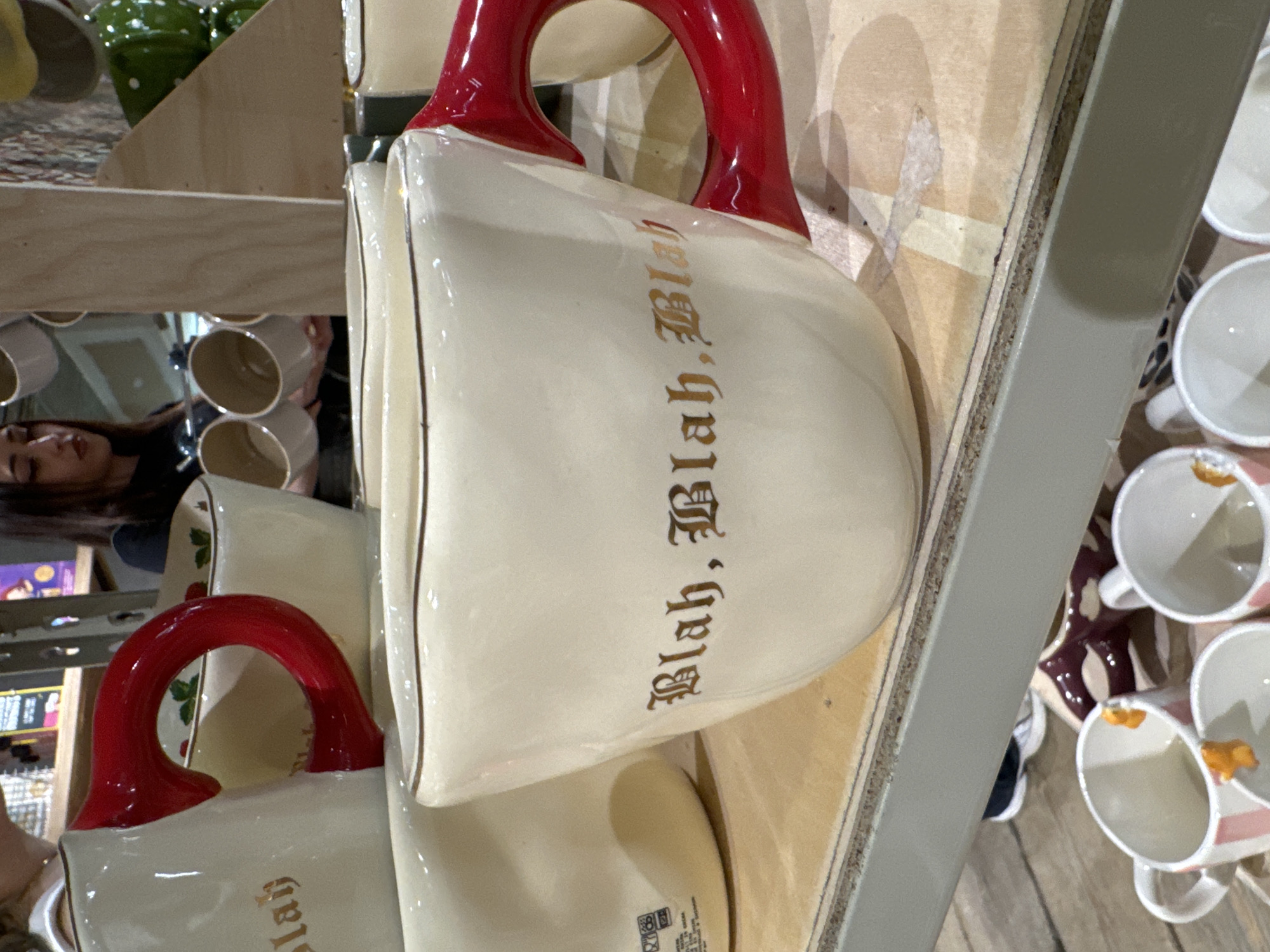 This is an indoor photograph of mugs on a shelf, likely in a retail setting. Two mugs, predominantly white with red handles, are the most prominent objects. They feature a decorative, old-style font displaying the text "Blah, Blah, Blah". Other mugs of varying designs and colors are visible in the background, suggesting a store or market. In the background, a reflection can be seen of a person, possibly a shopper in the store. This is likely a store that sells novelty items. This image does not allow me to confirm that this is in Barcelona, Spain.