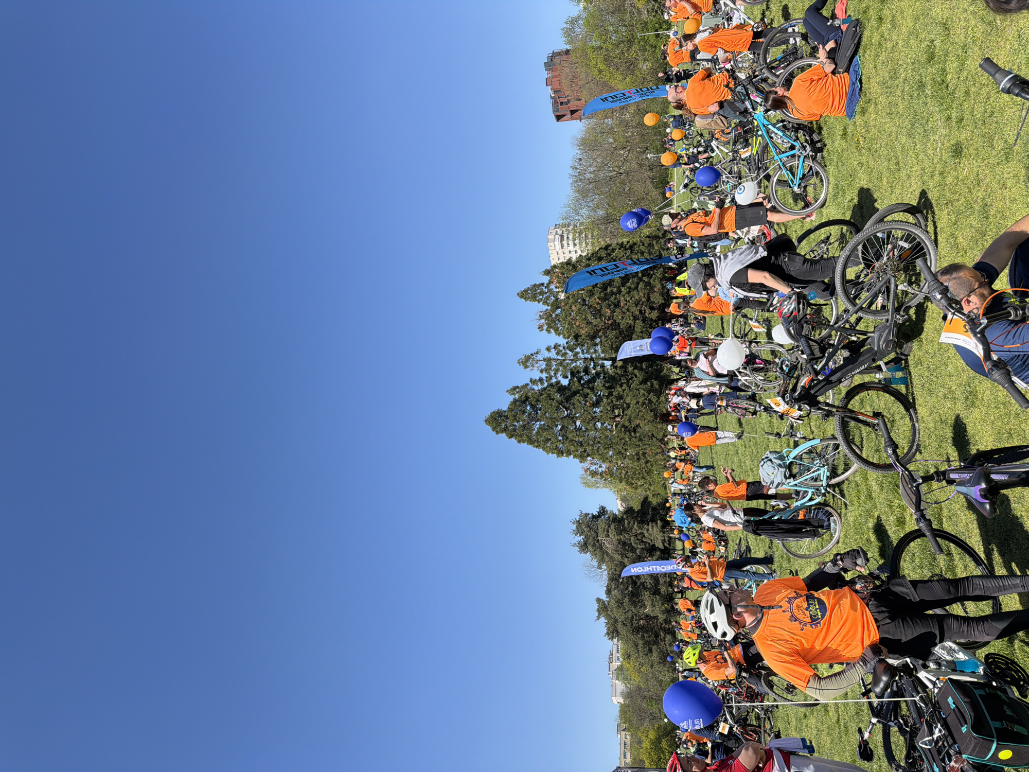 The image shows a large group of people and bicycles gathered on a grassy area under a clear blue sky, likely in a park. Many people are wearing orange shirts, suggesting a coordinated event or gathering. Several blue balloons and banners with the text "DECATHLON" are also visible. The presence of buildings and trees in the background indicates an urban environment. Given the location, the gathering is likely in a park within the city of Sofia, Bulgaria. People are resting on the grass near their bicycles, indicating a break or the end of a cycling activity.