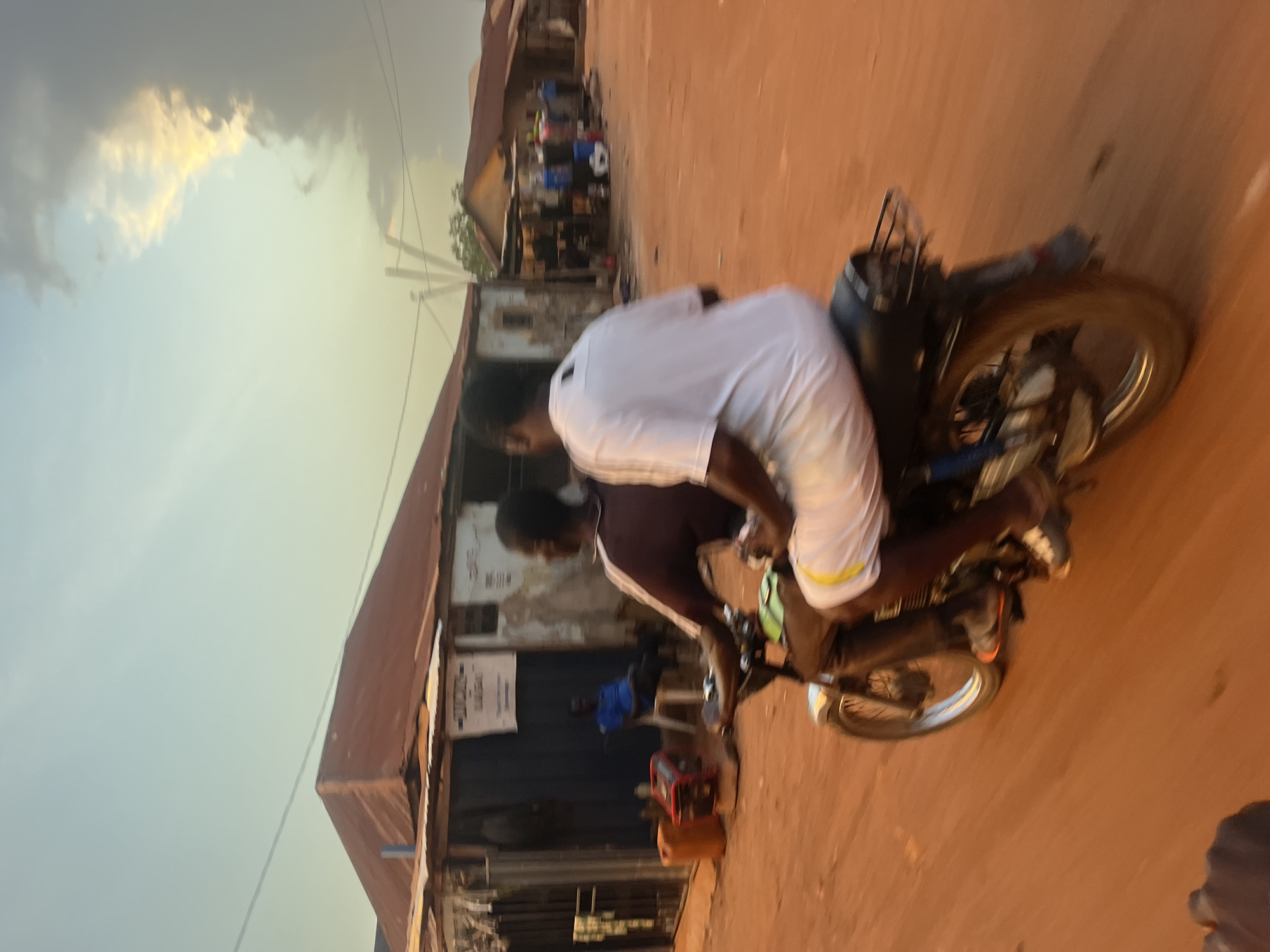 The image depicts two individuals on a motorcycle moving along a dirt road in a densely populated area. One person is driving while the other is a passenger. The environment shows rudimentary infrastructure, including buildings with rusted metal roofs, power lines, and dirt roads. The buildings have signs posted on them. The overall atmosphere suggests a scene in a lower-income, urban area. There is a generator and a jerry can on the side of the road. It is difficult to determine the precise location in Abuja, Nigeria.