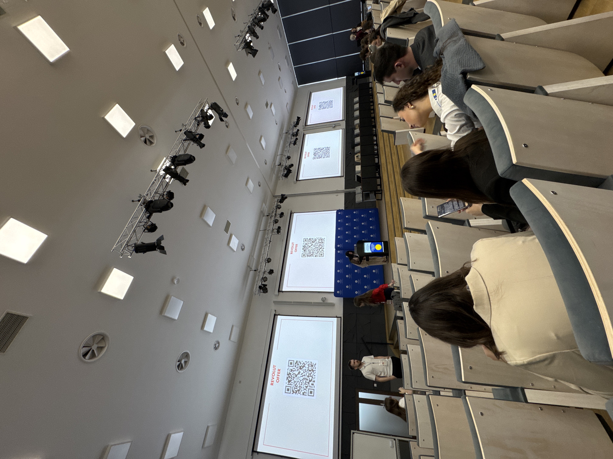 A photograph shows an audience in an auditorium. Several people are seated in rows of wooden chairs with blue padding. Some are looking at screens with QR codes labeled "REVOLUT OFFER". A stage is visible, with a person speaking near a blue backdrop. The location is in Bydgoszcz, Poland.