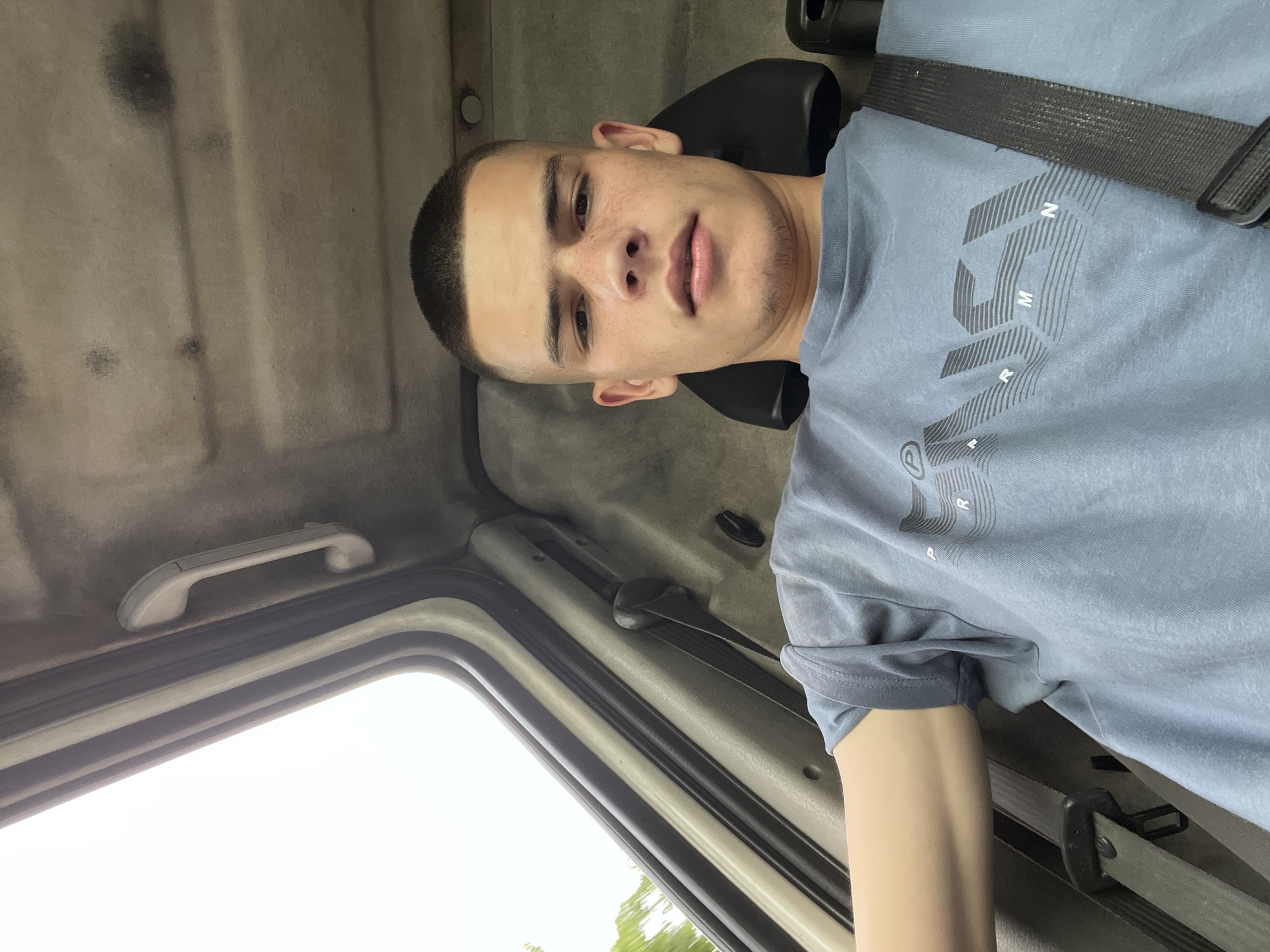 A young man with a buzz cut and light skin is sitting in what appears to be the driver's seat of a vehicle. He is wearing a light blue t-shirt with a graphic design on it. The t-shirt has stylized letters that are partially visible, including "M", "R", and "P". He is wearing a black seatbelt over his shoulder. The background suggests the interior of a truck or van, with a fabric-covered ceiling and a gray handle on the left side. The ceiling has some darker smudges and appears slightly worn. A portion of the window is visible on the left, showing an overcast sky and some greenery outside, suggesting it might be daytime and cloudy. The man's expression is neutral. The overall impression is a candid, slightly angled selfie taken inside a vehicle in Iganovo, Bulgaria.