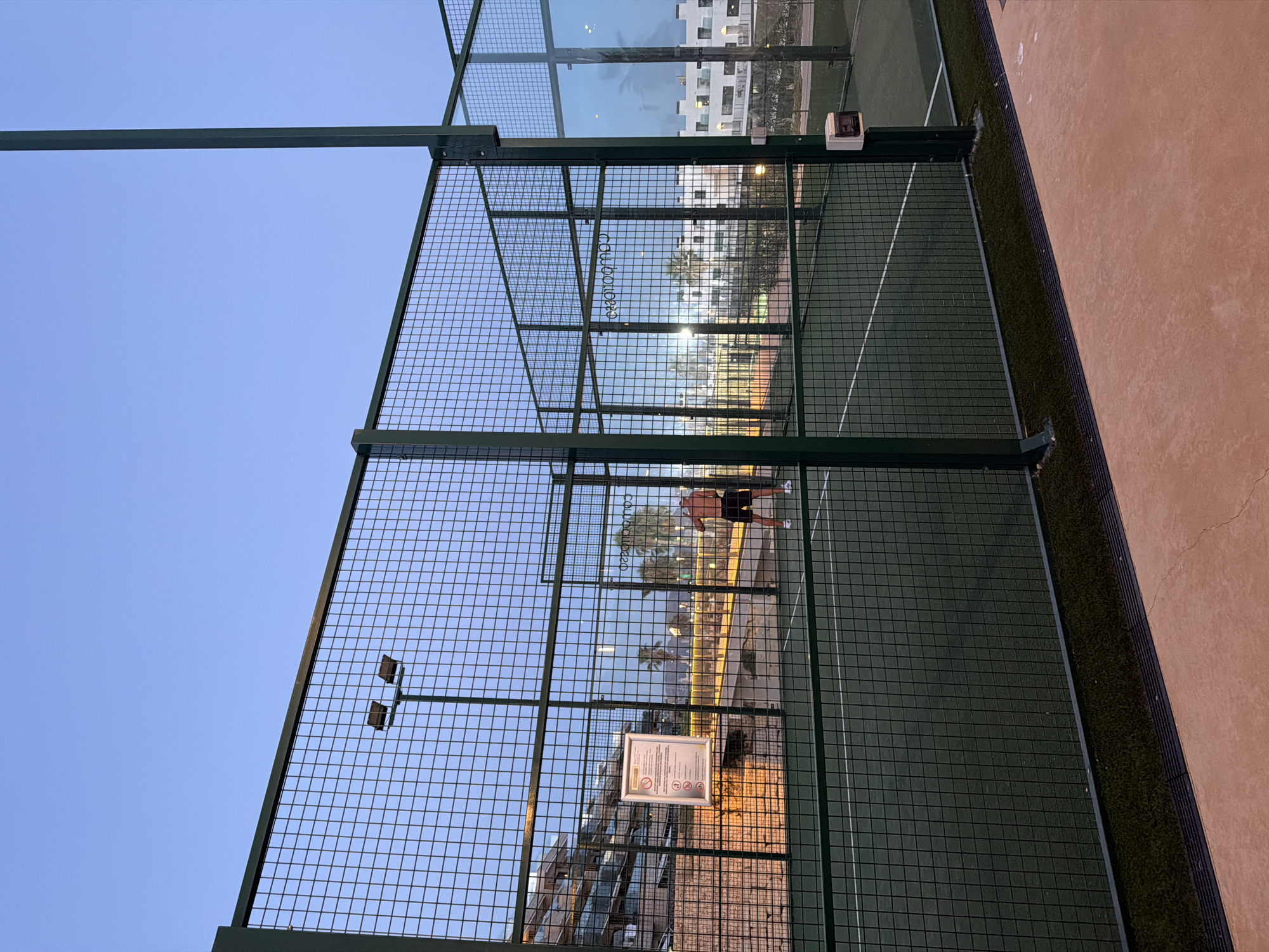 An outdoor padel tennis court is depicted in the city of Finestrat, Spain, at dusk. The court features a green artificial turf surface with white boundary lines and is enclosed by a green metal mesh fence. A man, shirtless and wearing black shorts and white shoes, is the sole visible person on the court, in the process of playing. In the background, beyond the fence, there are buildings, palm trees, and lights. A sign is attached to the fence on the left side of the image. The sky is a clear, pale blue, indicating the transition from daylight to evening.