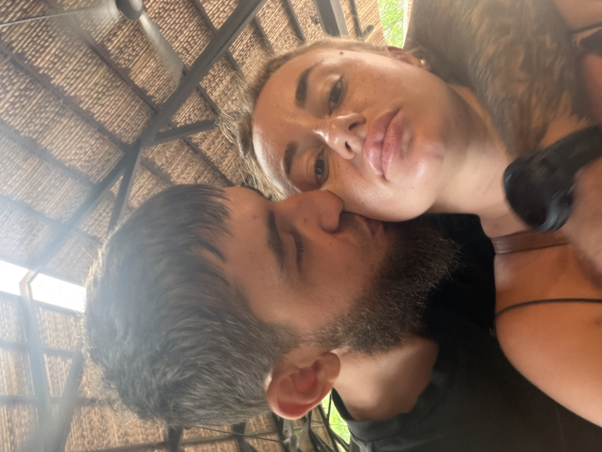 A man with a beard is kissing a woman on the cheek. They are indoors, with a thatched roof above them. A ceiling fan is visible in the background. The woman has freckles and full lips. The man is wearing a black t-shirt. The scene appears to be in Nha Trang, Vietnam.