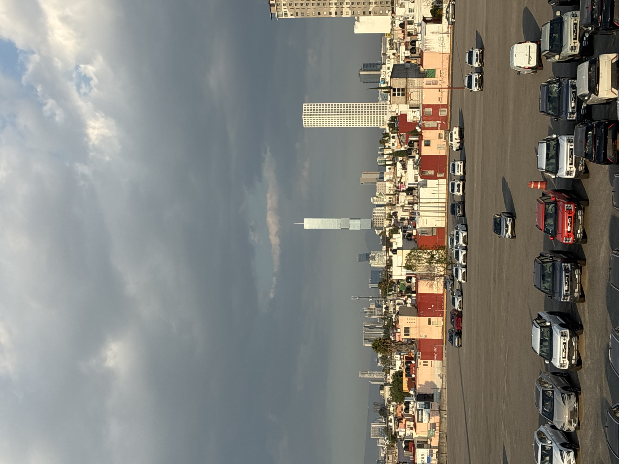 The image depicts an elevated view in San Bernardino Tlaxcalancingo, Mexico. In the foreground, a large, paved lot contains rows of parked vehicles. The vehicles appear to be primarily trucks. Adjacent to the lot, a dense urban area consists of low to medium-rise buildings with varied architectural styles and color schemes. In the distance, a skyline is punctuated by taller buildings and skyscrapers. The sky above the scene is filled with grey, cloudy formations, indicating potential inclement weather. There are no visible people in the image.