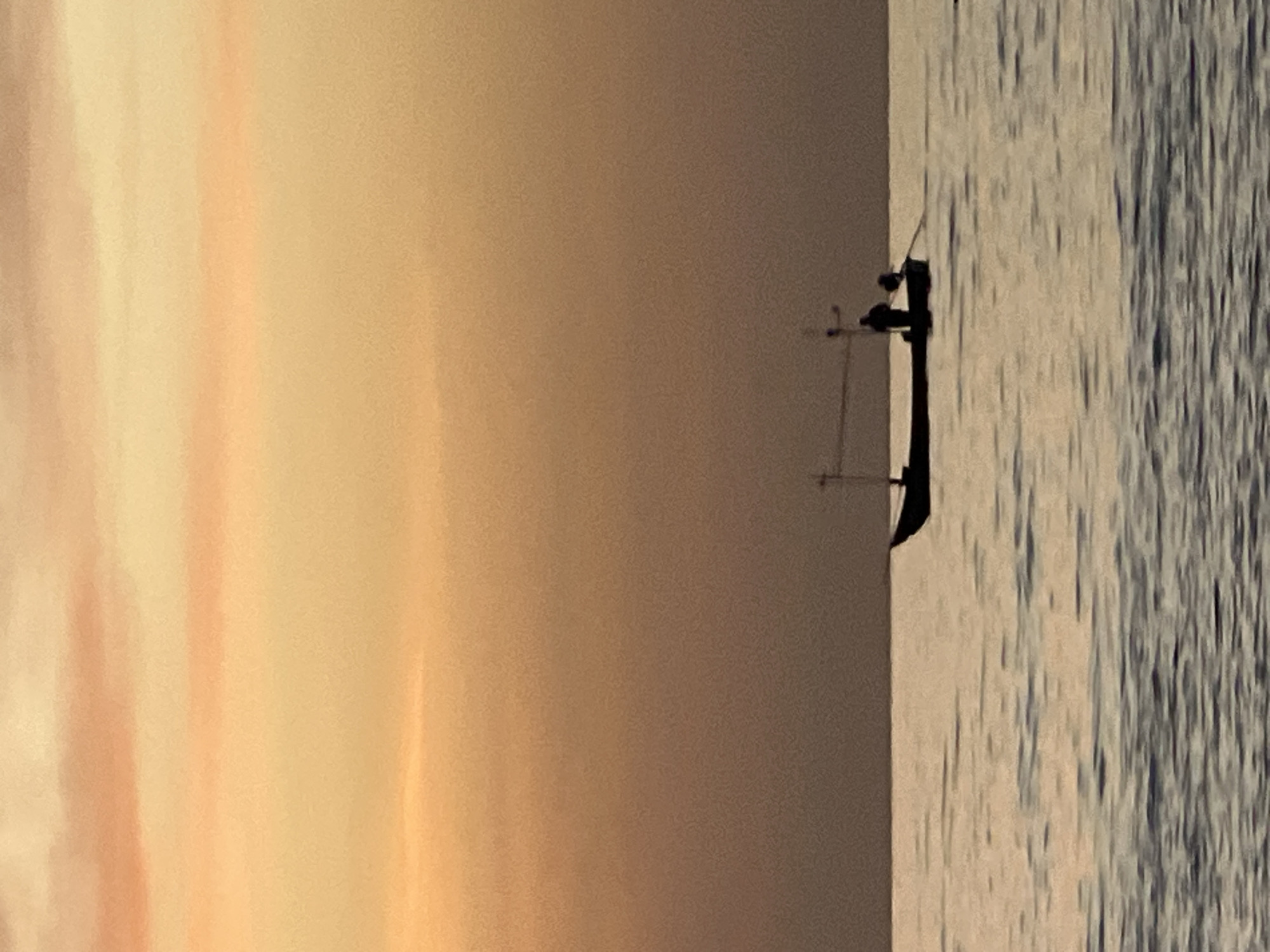 A silhouette of a fishing boat with one person on board is visible on calm waters. The boat has a single mast and a thin railing along its side. The person appears to be holding an oar. The water is slightly rippled, reflecting the soft colors of the sky. The sky is a gradient of warm hues, transitioning from a soft orange and pink at the top to a muted brown near the horizon, suggesting either sunrise or sunset. The scene takes place in the sea off the coast of Ko Pha-ngan District, Thailand.