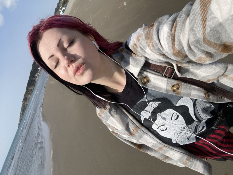 The image displays a young adult female captured in a high-angle selfie on a sandy beach in Ulcinj, Montenegro. She has closed eyes and puckered lips, facing slightly right of center. Her hair is styled with a prominent split dye, dark on the left side and magenta on the right. White wired earphones are inserted into both ears, with the cord draped across her upper chest. She is wearing a grey, white, and brown plaid shirt-jacket, unbuttoned, over a black t-shirt featuring a distressed white and grey graphic print of a stylized female face holding an apple. A brown leather strap from a cross-body bag is visible over her right shoulder. Partially visible below her t-shirt are red and black plaid trousers or skirt. The immediate foreground consists of dark, wet sand with gentle ocean waves receding. In the background, the sea extends to a distant coastline marked by light-colored cliffs and hills under a clear blue sky. Faint white structures are visible on the far right along the distant shoreline.