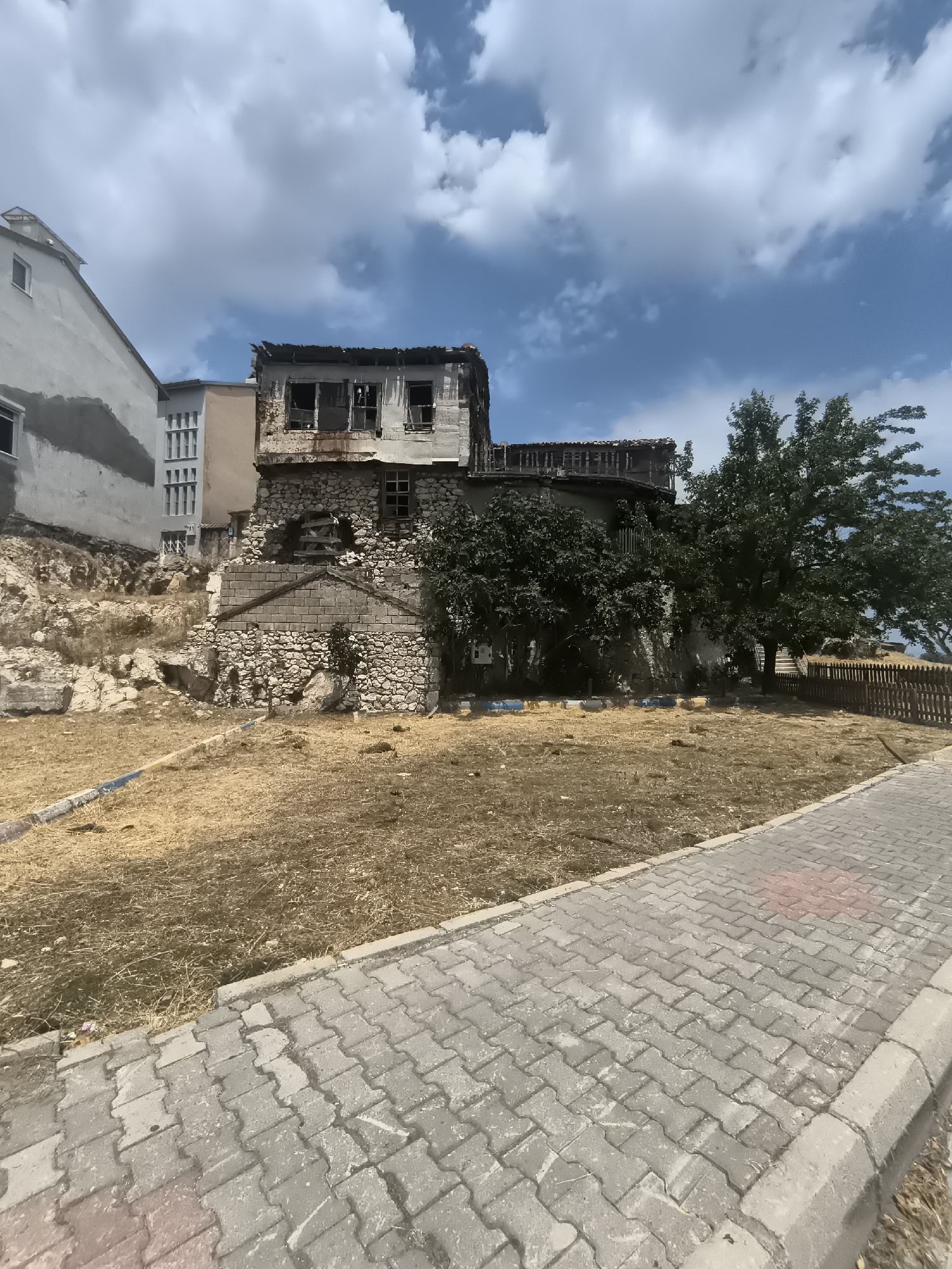The image depicts an abandoned, multi-story building with a stone foundation and weathered wooden upper floors, situated in a dry, grassy area. The building appears structurally unsound, with damaged roofing and boarded-up windows. Adjacent to it, a modern, multi-story building with a light-colored facade and visible windows stands on a higher elevation. The foreground shows a paved walkway with interlocking gray pavers, bordered by a curb. The background includes a large, mature tree, a wooden fence, and a blue sky with scattered clouds. A portion of the area around the abandoned building is covered in dry, straw-like vegetation. The scene appears to be in a semi-urban or historical district. The location is identified as Eğirdir, Turkey.