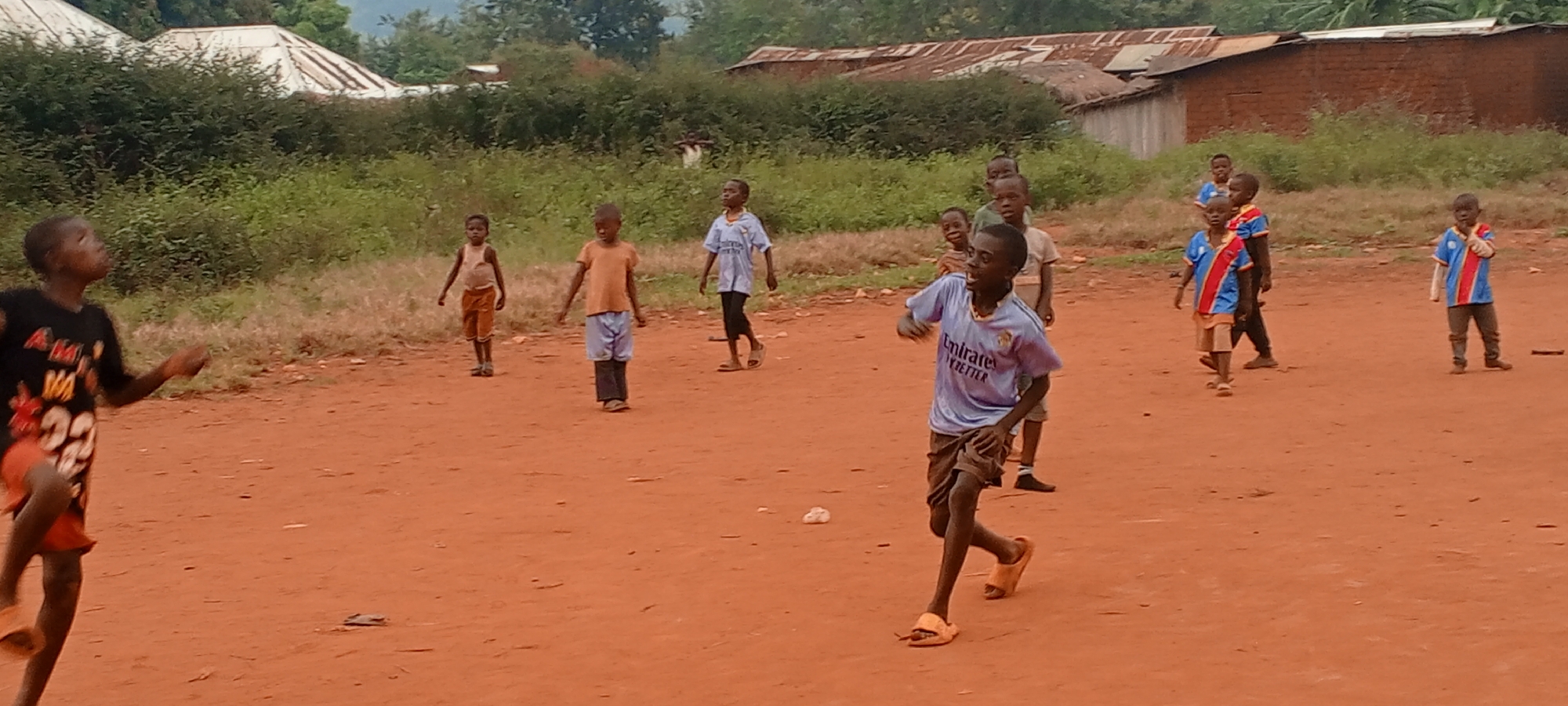 The image depicts a group of approximately ten children on a reddish-brown dirt field in what appears to be an outdoor setting. They are of African descent, and most are wearing casual clothing, including t-shirts, shorts, and sandals. A few are wearing matching blue, red, and white striped shirts. They seem to be engaged in play, with one child in the foreground running.
In the background, there is a line of dense green vegetation, behind which several small buildings with corrugated metal roofs can be seen. The overall impression is that this is a rural or semi-rural area. The scene is likely in Ngalula, Democratic Republic of the Congo, based on the prompt's information.