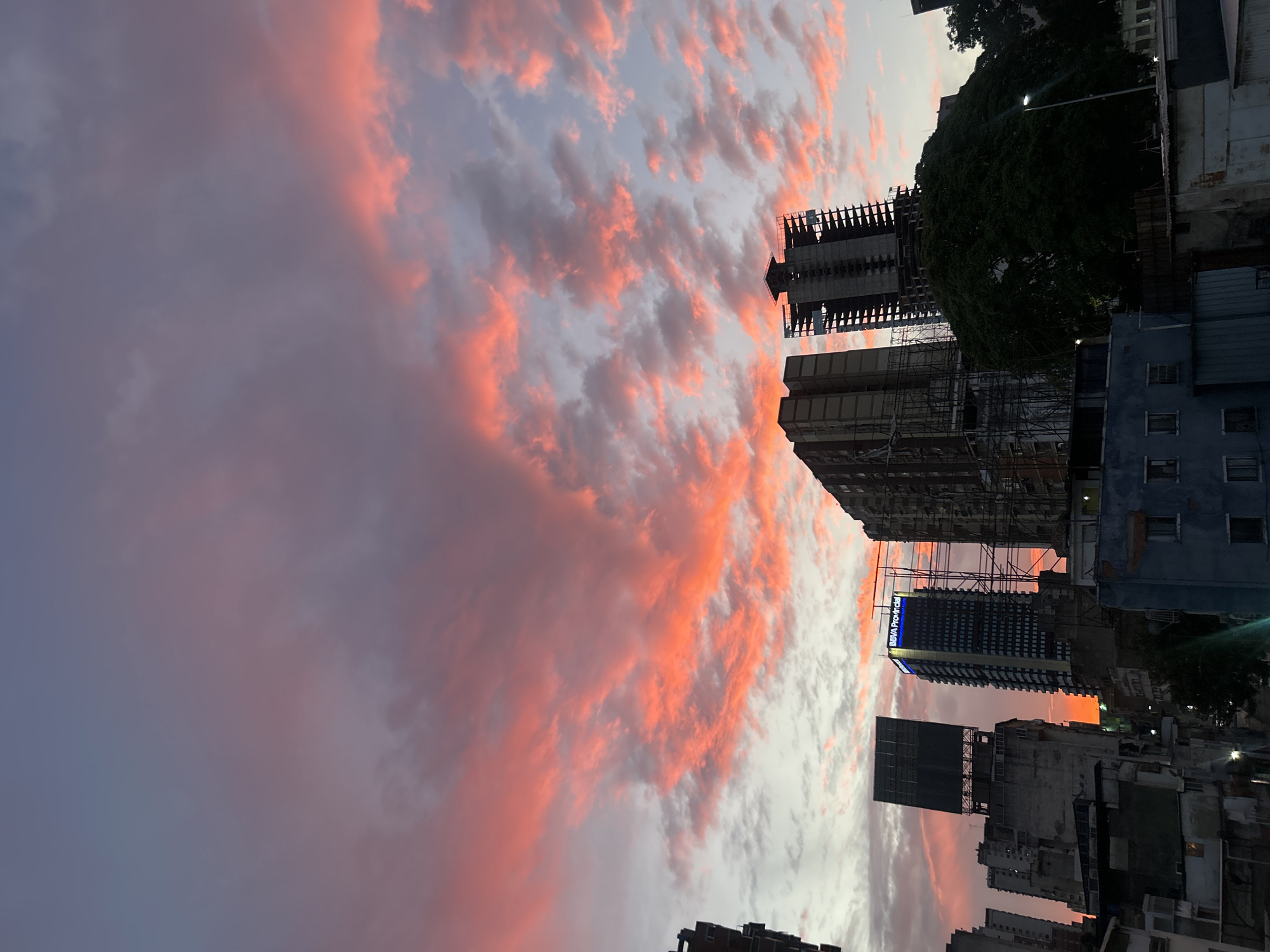 The image depicts a cityscape at sunset in Caracas, Venezuela. A vibrant pink and orange sky dominates the upper portion of the frame, with clouds illuminated by the setting sun. In the lower portion, buildings of varying heights are visible, interspersed with trees. Construction scaffolding is present on some of the taller buildings. A building is branded “BBVA Provincial”, a local Venezuelan Bank.