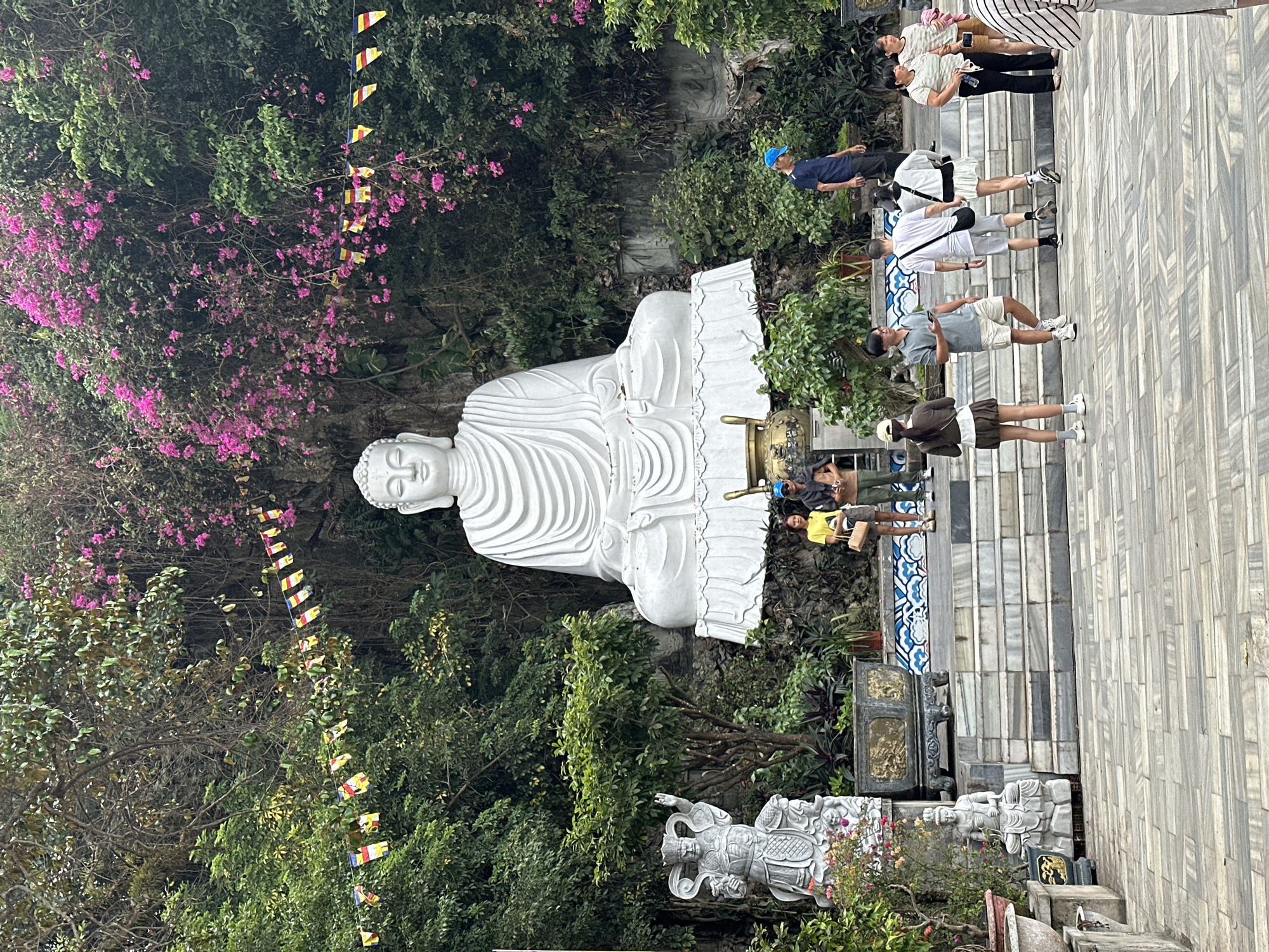 This is a photograph taken at the Marble Mountains in Đà Nẵng, Vietnam. The central subject is a large, white, seated Buddha statue situated amidst lush green foliage and pink flowering bushes. In front of the Buddha, there are steps leading up to a platform. Several people are visible on these steps and in the foreground, some are looking at the statue, others are interacting with each other. To the left of the Buddha statue, there is a smaller, ornate stone sculpture of a warrior-like figure. Prayer flags are strung across the scene, adding to the spiritual atmosphere. The ground in the foreground is paved with grey and white patterned tiles.