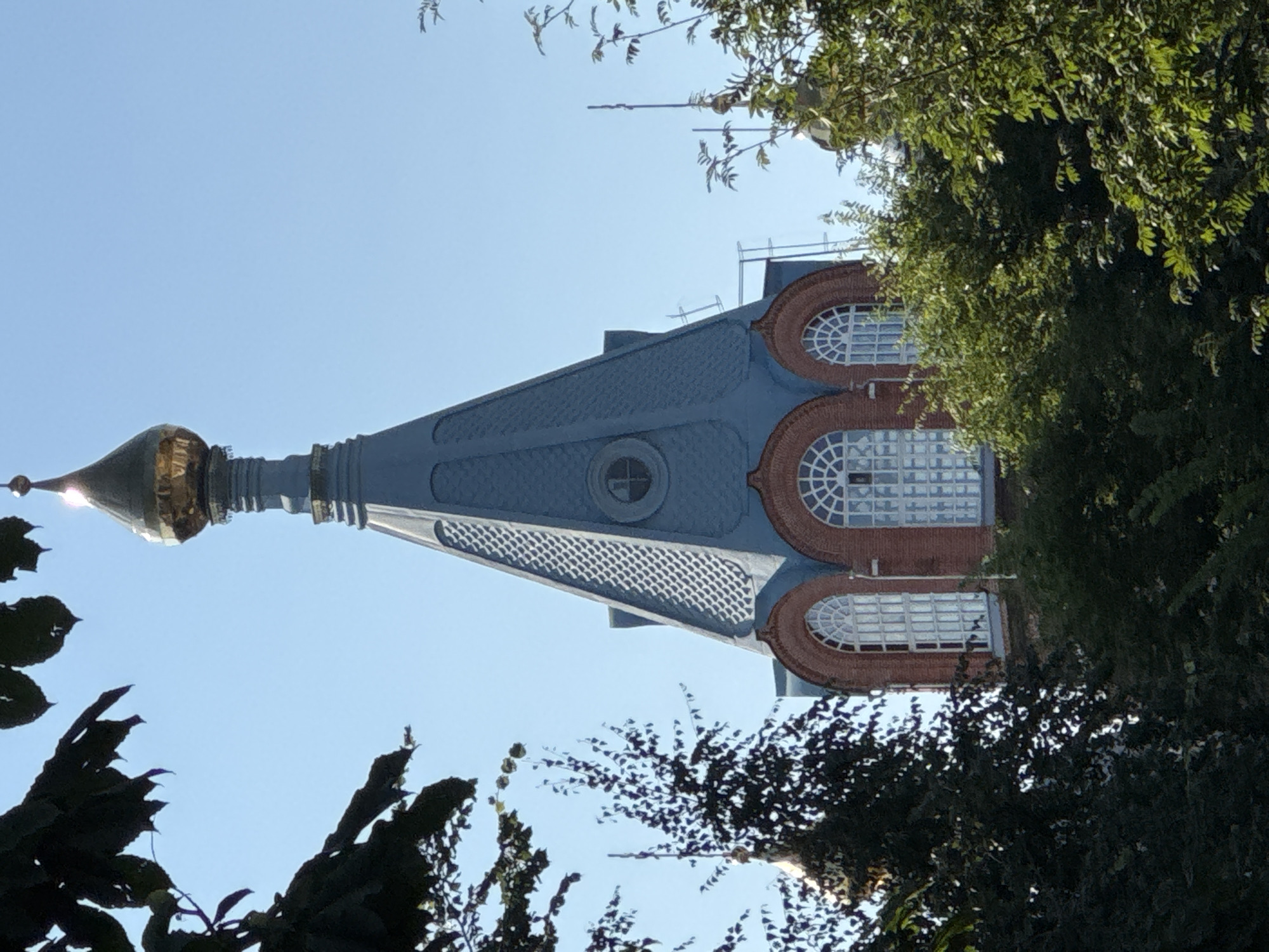 The image shows the top section of a church bell tower, characterized by its blue, conical roof topped with a golden dome. The tower has arched windows with intricate white lattice work, set within a red brick facade. A small circular window is visible higher up on the blue roof. Lush green trees partially obscure the lower parts of the tower and surround the structure, suggesting an outdoor setting. The sky is clear and blue, indicating daytime. The architectural style suggests it is likely a Russian Orthodox church, possibly located in the historic center of Nizhny Novgorod, Russia, known for its many churches and cathedrals.