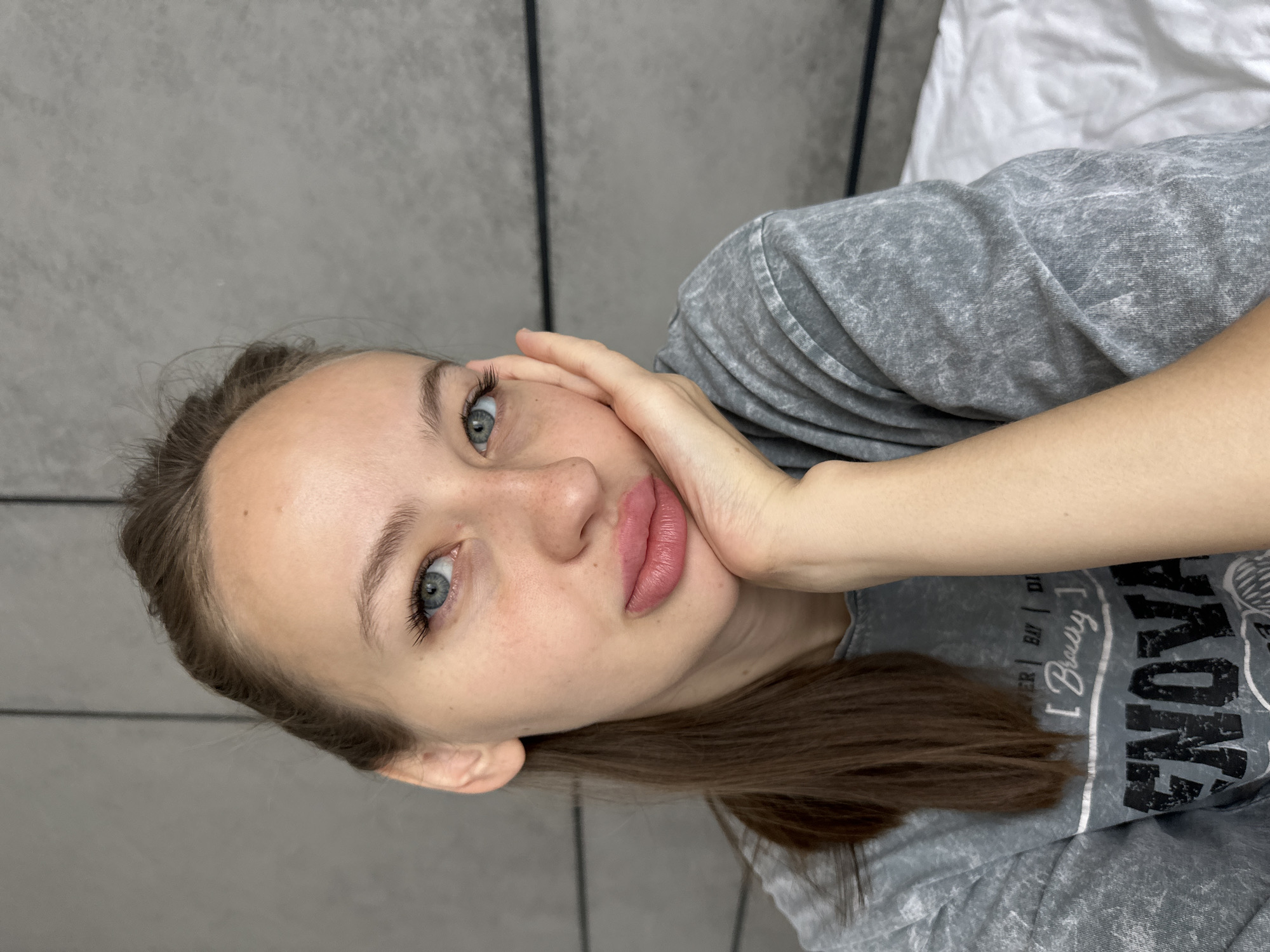 A young woman is lying down, her head resting on her hand. She has fair skin, blue eyes, and light brown hair. She is wearing a gray t-shirt with a graphic design. The background consists of a gray wall and a white surface. There is no indication of the location being in Đà Nẵng, Vietnam.
