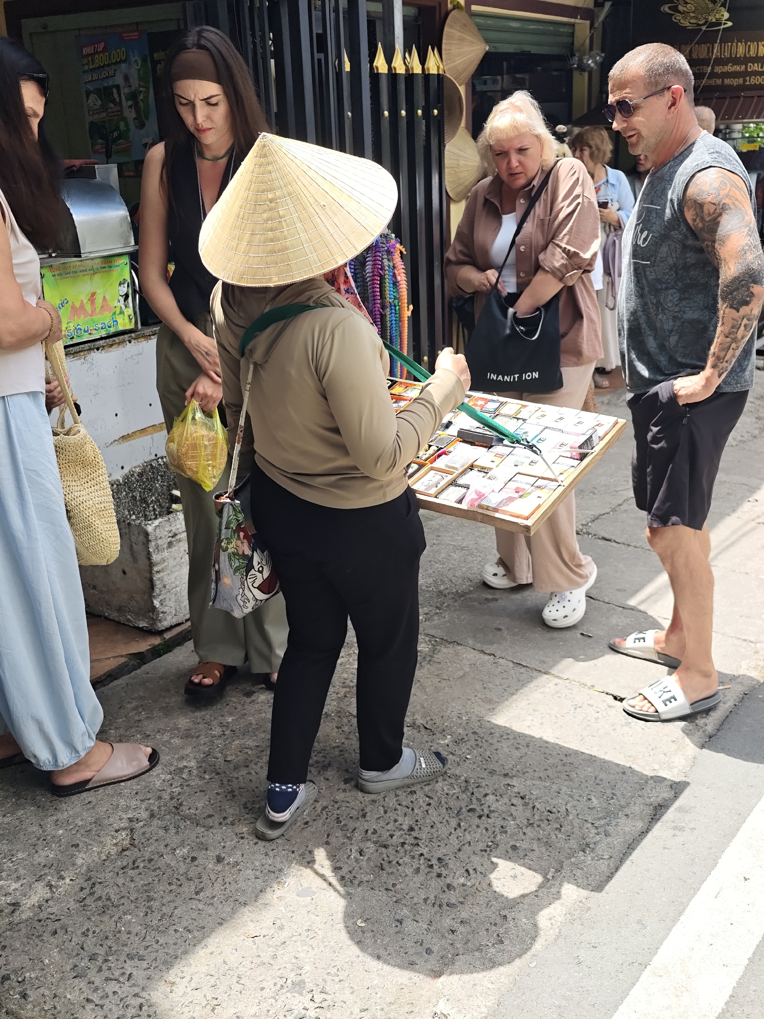 In Da Lat, Vietnam, a street vendor wearing a conical hat displays a tray of small trinkets. She is surrounded by four people. To her left, a woman in a white top and light blue skirt looks down at the wares. Next to her, another woman in a black vest and olive pants holds a yellow plastic bag containing items. To the vendor's right, a woman in a brown jacket and beige pants examines the items, holding a black tote bag. A man with tattoos and sunglasses stands closest to the vendor on the right. The vendor's tray is filled with various small, colorful items arranged in compartments. The background features a building with signage and decorative elements, suggesting a commercial area. The ground is paved with concrete.