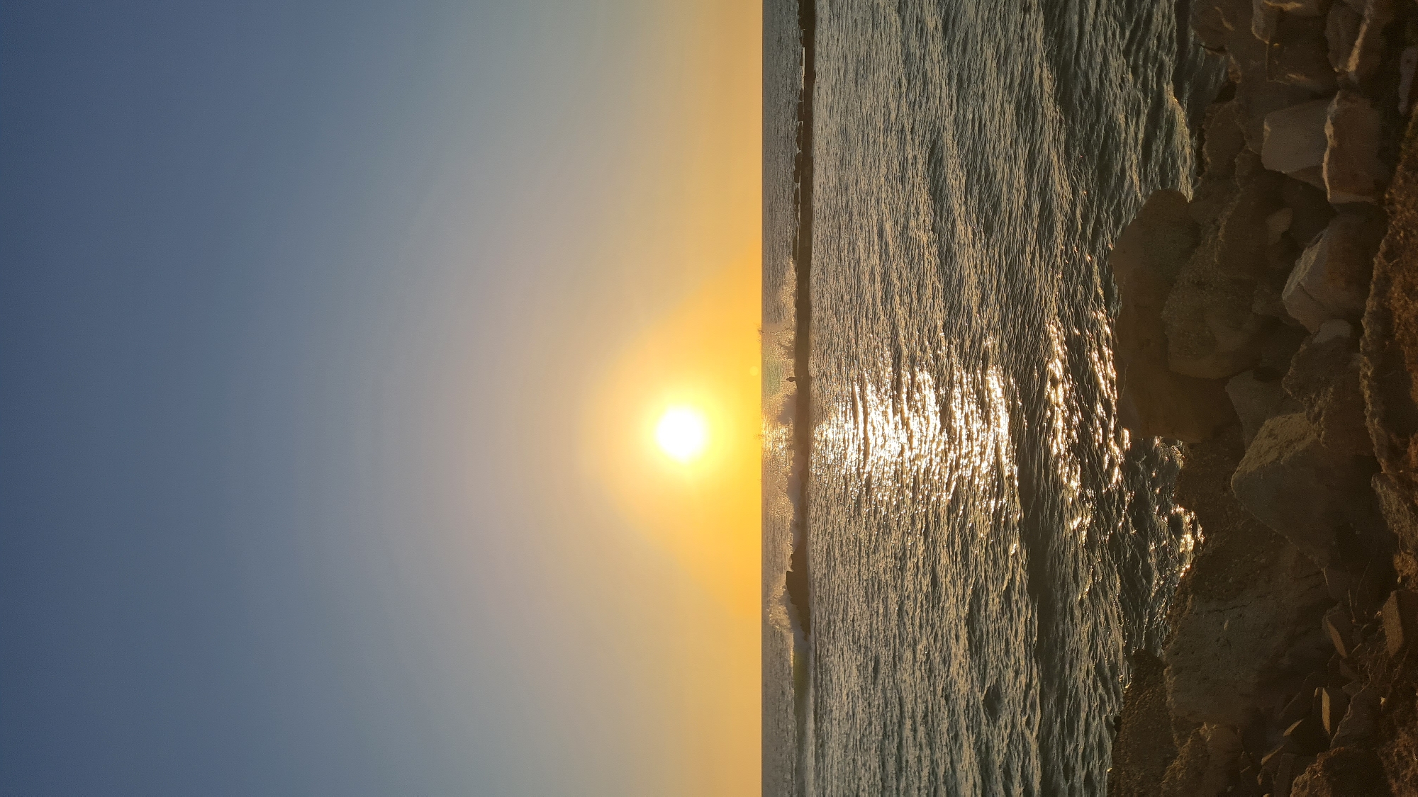 The image shows a sunset view in Nahariyya, Israel. The sun is positioned in the center, casting a bright light across the sky and reflecting on the sea. The sky transitions from blue to golden hues closer to the sun. The sea is on the right, with waves reflecting the sunlight. Rocky terrain occupies the lower right corner of the image.