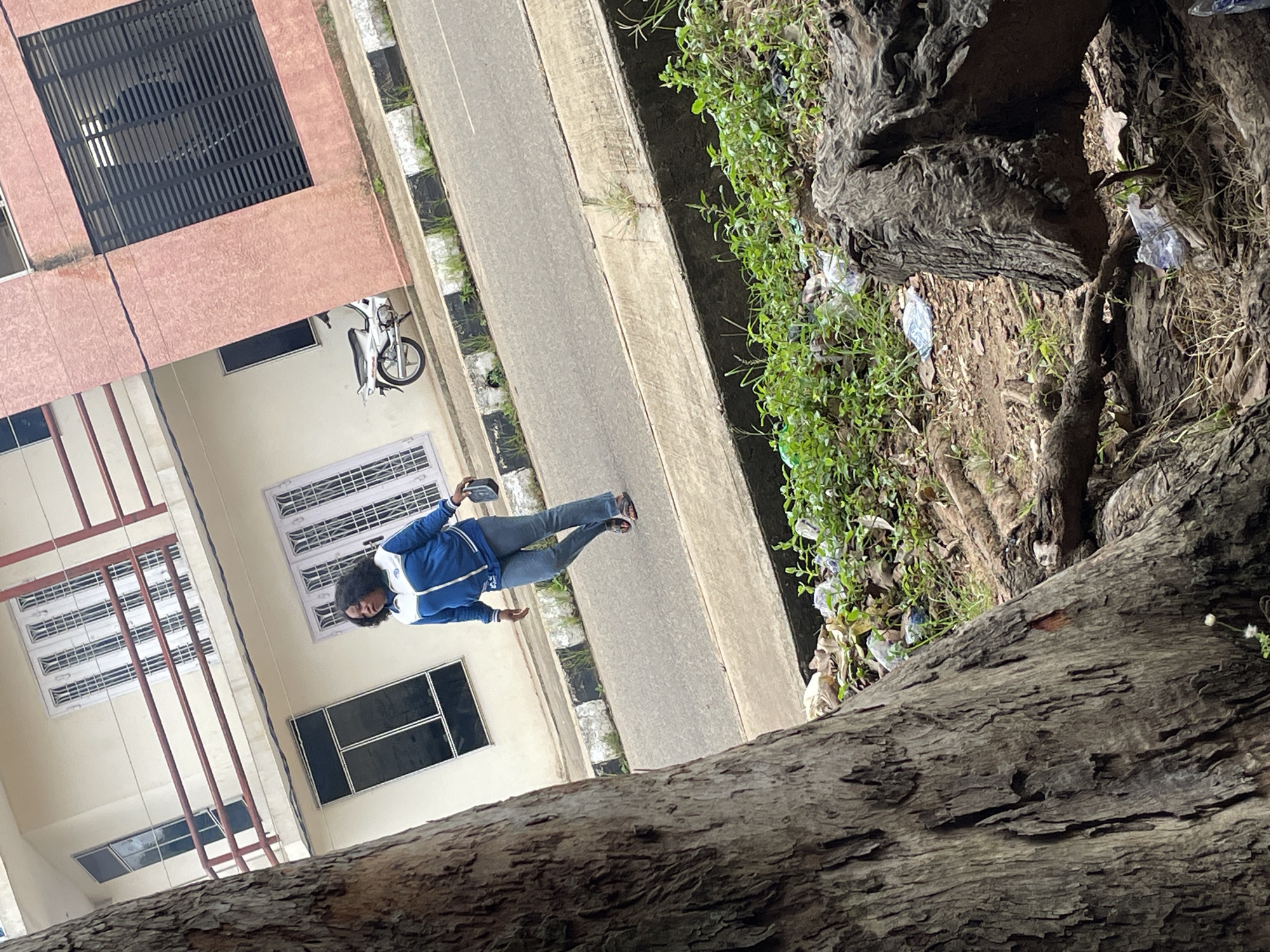In Jos, Nigeria, a woman in a blue and white jacket and jeans walks along a street. She is carrying a black object in her right hand. She is walking on the sidewalk in front of a light-colored building with a red railing and a pink building with black shutters. A white motorcycle is parked on the sidewalk in front of the pink building. The street is paved and has a concrete gutter. The foreground of the image shows the base of a large tree with exposed roots and some trash scattered on the ground.