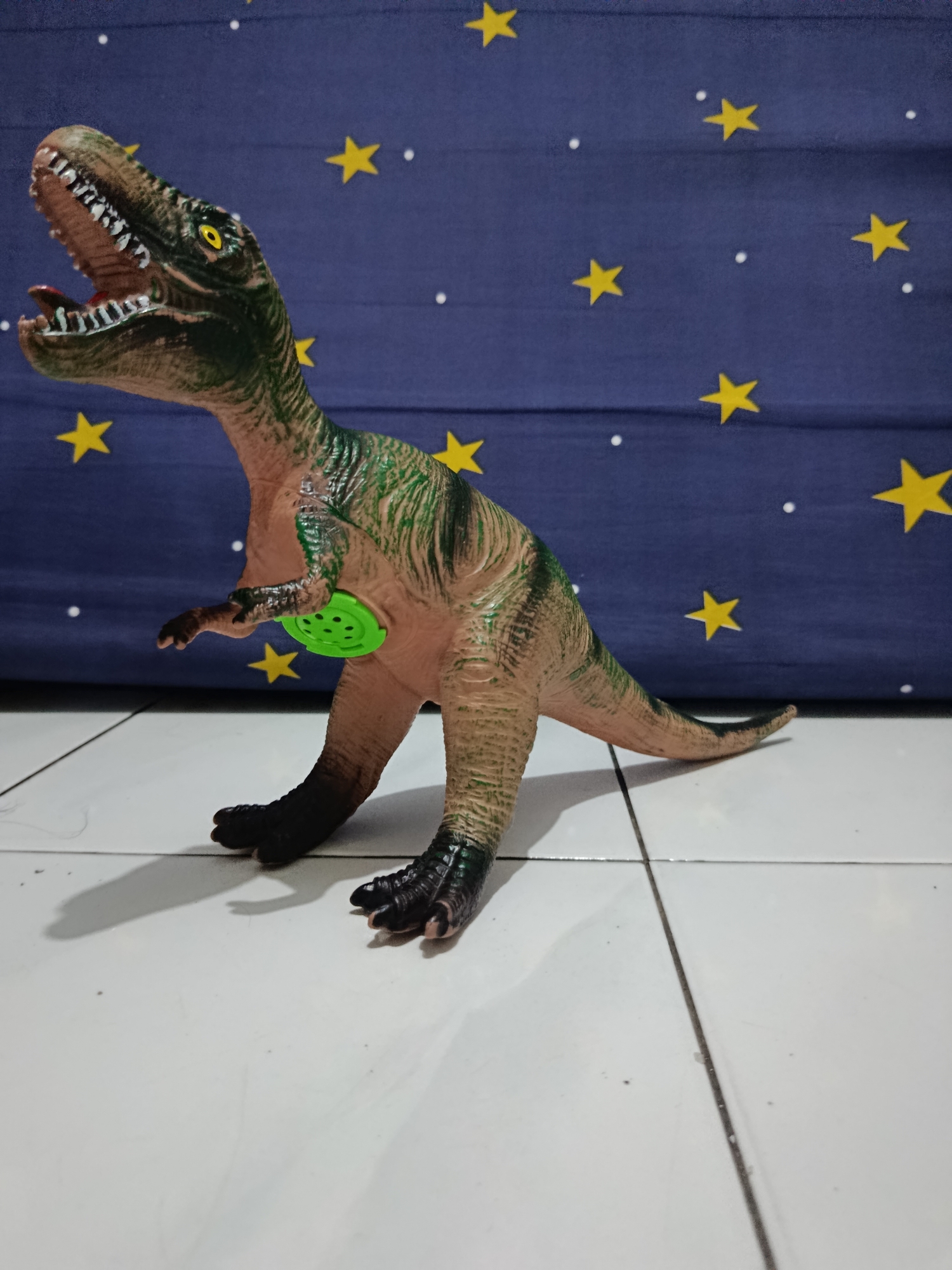 A toy Tyrannosaurus Rex dinosaur stands on a white tiled floor, its body brown and green with textured skin. Its mouth is open, revealing sharp white teeth and a red interior. The dinosaur has yellow eyes and small, underdeveloped forelimbs. A bright green circular object with perforations is attached to its underside. In the background, a dark blue fabric hangs, decorated with yellow stars and small white dots, resembling a night sky. The scene is lit from the front, casting a shadow of the dinosaur onto the floor. The image appears to be taken indoors, likely in a residential setting. No people are present in the image. The location of Wanasari, Indonesia is not ascertainable from the visual elements of the image.