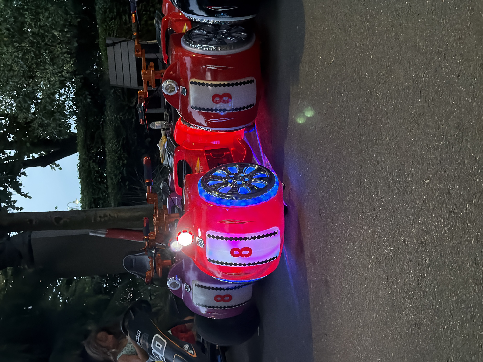 A low-angle shot shows a row of brightly colored, miniature race cars lined up on pavement. The cars, lit with vibrant red and purple LEDs, have a design reminiscent of classic F1 racers, complete with racing numbers "8" on their sides. One car is a deep red, another is a vibrant purple, and a third is black. The wheels of the red car are particularly striking, featuring illuminated blue lights. In the background, a young girl with light brown hair, wearing a light-colored top, is visible to the left, seemingly interacting with one of the cars. Behind her, a dark, somewhat indistinct structure stands tall, possibly part of the urban landscape. Trees and hedges form a dense green backdrop, suggesting an outdoor setting in a park or recreational area. The sky above is a pale blue, indicating it is likely late afternoon or early evening. The pavement appears slightly wet or reflective. The overall atmosphere is playful and energetic, with the illuminated cars creating a futuristic and captivating display. This scene is likely located in the city of Burgas, Bulgaria, specifically in the area near the Sea Garden, known for its recreational attractions and public spaces, which often feature similar attractions for children.