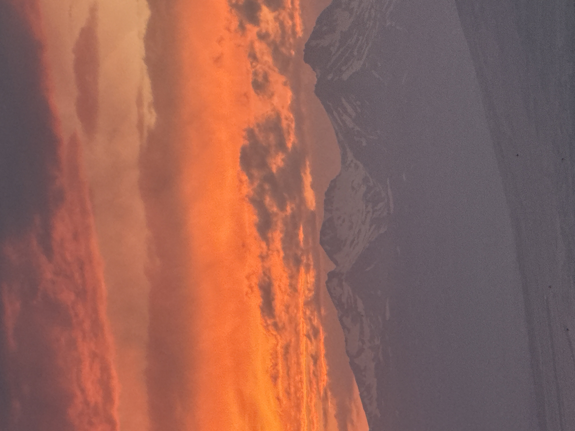 A mountainous landscape is depicted under a vibrant, fiery sunset. The sky is ablaze with hues of orange and red, transitioning to darker purples at the top, with scattered clouds adding texture to the dramatic sky. In the middle ground, a range of snow-capped mountains forms a silhouetted, imposing backdrop. The foreground is a gentle, snow-covered slope, its surface muted by the fading light and distance, obscuring any finer details. The scene is devoid of any human presence, structures, or identifiable landmarks that would pinpoint a specific location within Abovyan, Armenia, or any other city. The overall impression is one of natural grandeur and solitude.