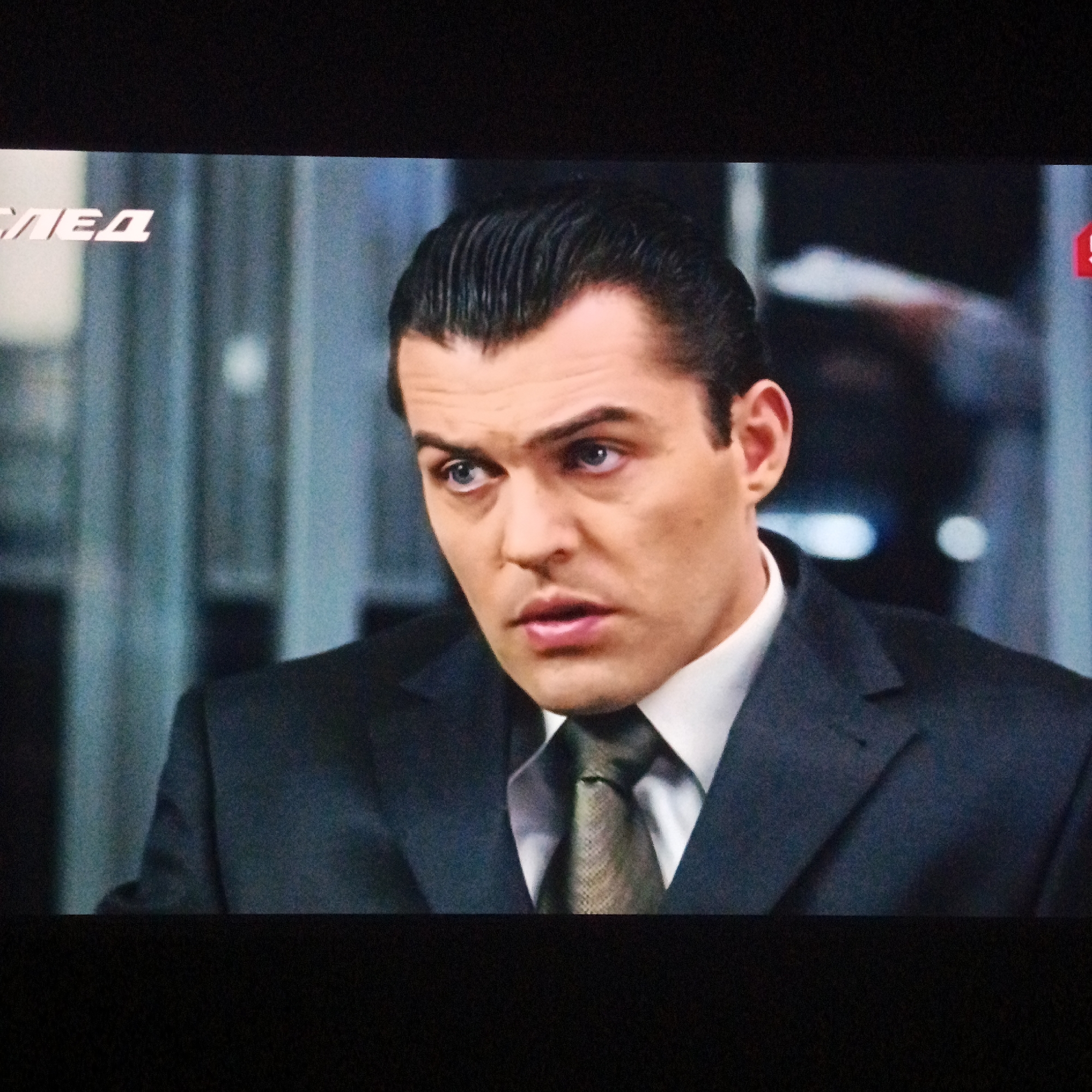 The image is a close-up shot of a man in a suit and tie. He has dark hair slicked back, light eyes, and a concerned expression. The background is blurred, showing what appears to be an office environment. A partial logo "СЛЕД" is visible in the top left corner, and part of the number "5" is in the top right. No location information is discernible from the image.