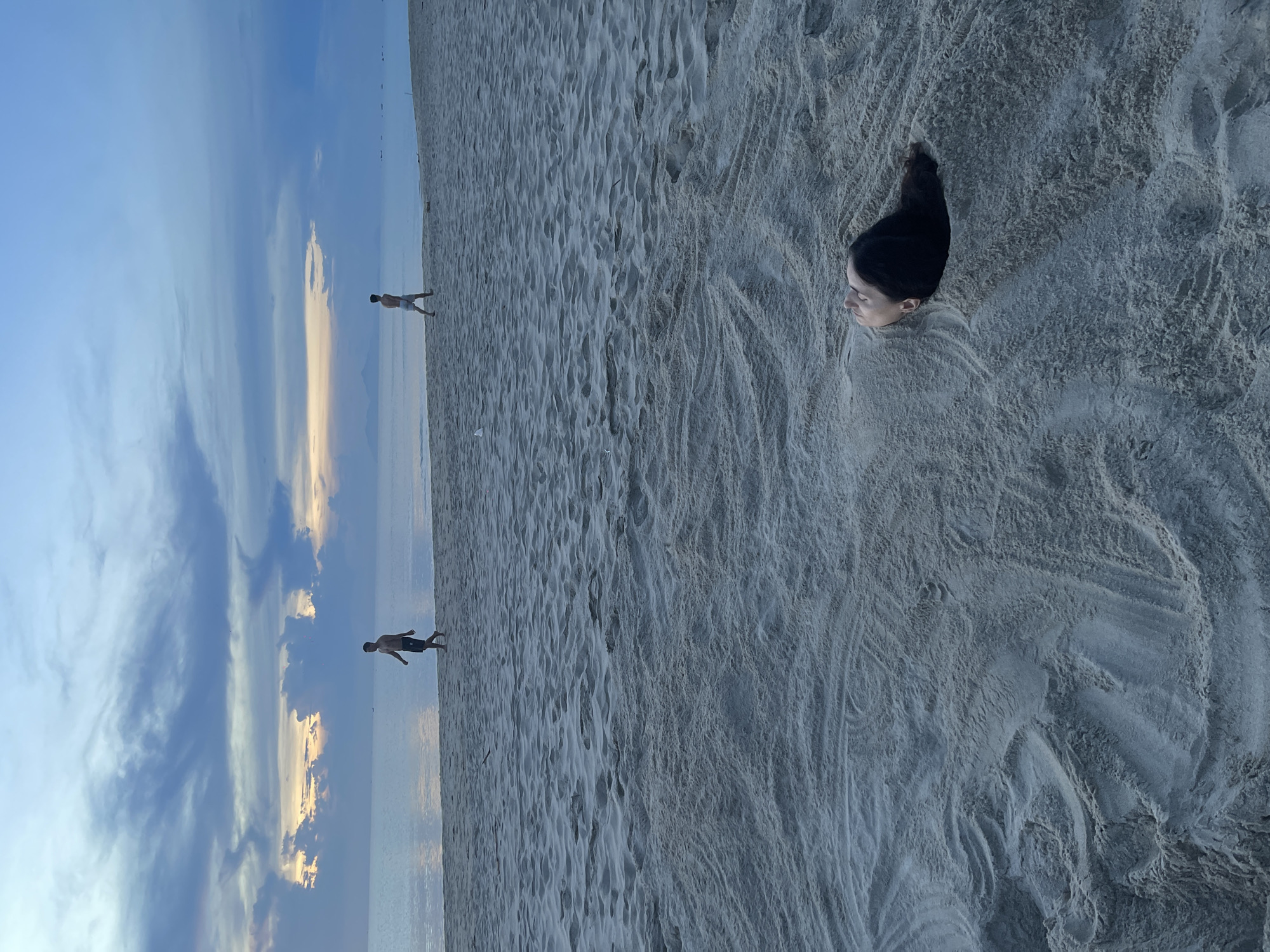 The image depicts a sandy beach scene at dusk, likely in Da Nang, Vietnam. In the foreground, a person's head and shoulders are buried in the sand, with their eyes closed and face turned upwards. The person has dark hair. The sand is textured, showing patterns from the tide or wind. Further back on the beach, two young males are walking away from the camera towards the water. They are silhouetted against the horizon. The ocean stretches to the horizon, reflecting the soft, diffused light of the setting sun, which is partially obscured by dramatic clouds in shades of blue and grey, with hints of orange and yellow peeking through. The overall atmosphere is tranquil and serene.