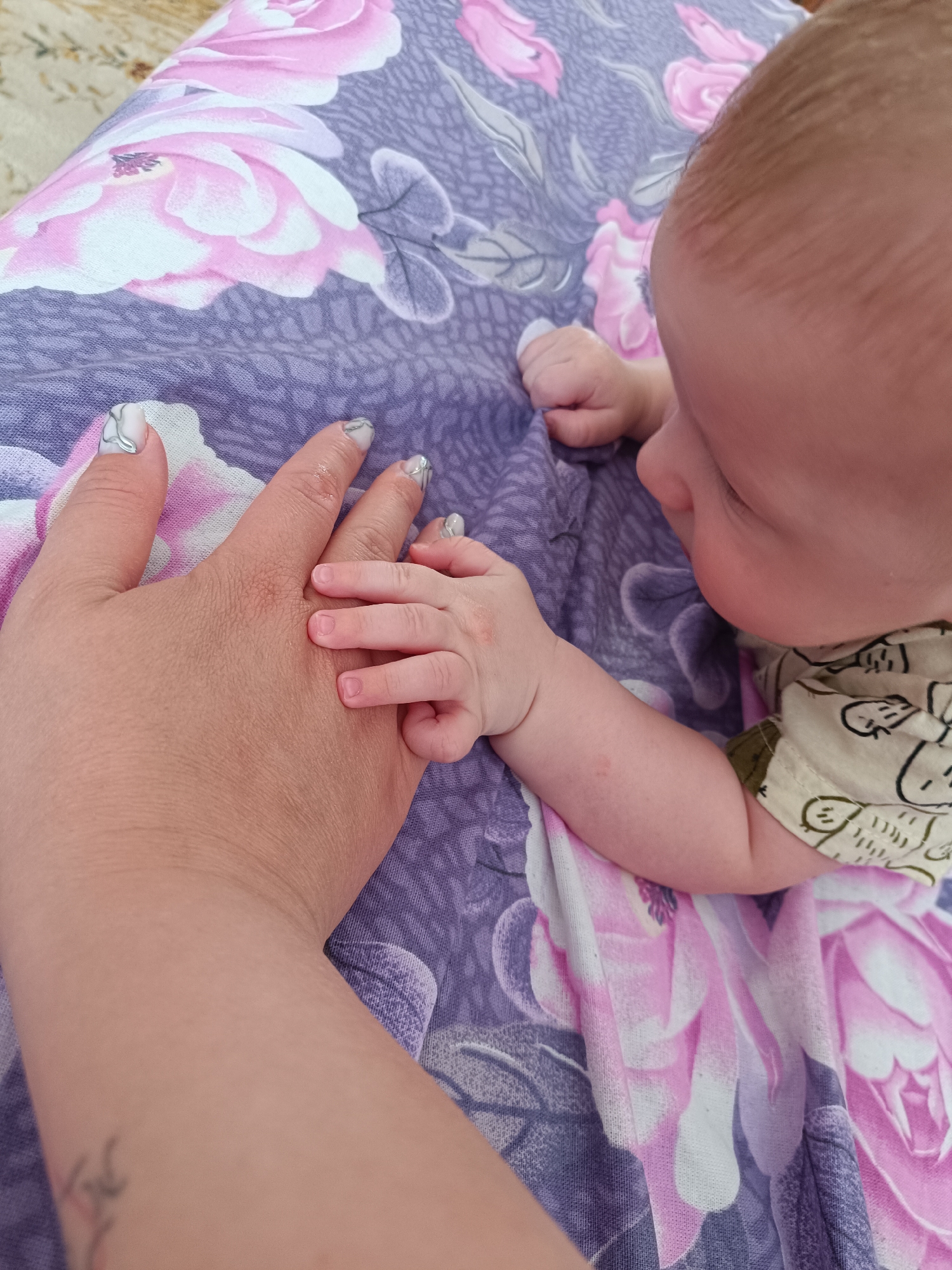 An overhead shot shows a baby lying on a patterned blanket, clutching the hand of an adult. The blanket has a repeating design of large pink and white flowers on a purple background. The baby has fair skin and blond hair, wearing a light-colored shirt patterned with dark, stylized animals. The baby's fingers are intertwined with the adult's fingers, who is out of focus, only showing their hand, with painted nails in light grey. A small tattoo is visible on the wrist of the adult. The location is unknown.