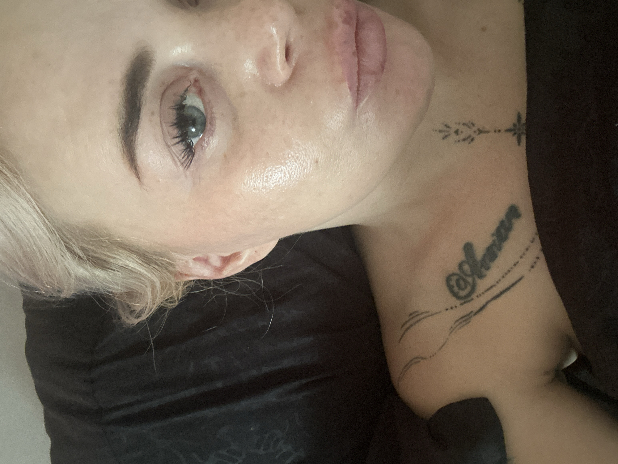 A close-up, high-angle, indoor shot shows the upper chest, neck, and right side of the face of a blonde woman lying down. The woman is looking towards the camera and her face is slightly angled away. She has light-colored skin with some freckles, light blonde hair, and blue eyes. Her lips are slightly parted and appear to be tinted pink. Her right eyebrow is dark and arched. She is wearing makeup, including mascara and eyeliner. She has several tattoos visible: one on her upper chest that appears to say "Amara" in cursive, and a smaller, more intricate tattoo on her neck that resembles a stylized floral or geometric pattern. She is wearing a dark-colored top that is partially visible. The background is a dark, textured fabric that appears to be a blanket or pillow. The lighting is soft and diffused, suggesting an indoor setting. No specific location within a city is discernible.
