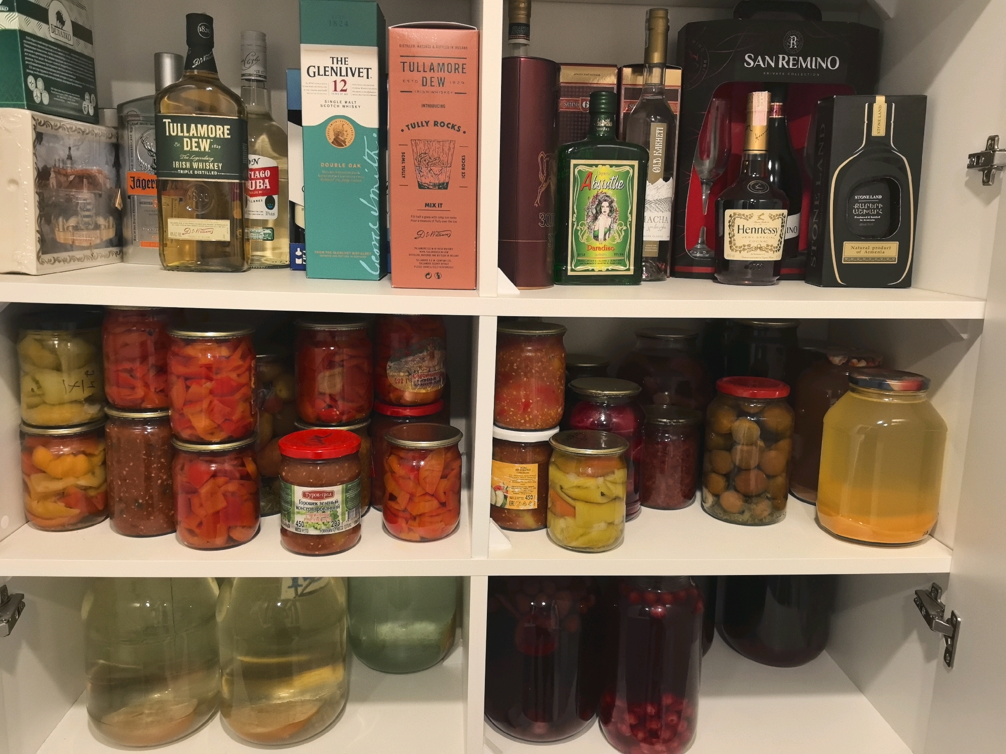 The image shows a pantry shelf stocked with various alcoholic beverages and preserved food items. On the top shelf, several bottles of whiskey and other spirits are visible, including a bottle of Tullamore Dew Irish Whiskey and a bottle of The Glenlivet 12 Year Old Scotch Whisky. There are also jars of preserved peppers and other vegetables on the middle shelf. The bottom shelf contains large glass jars with what appear to be pickled fruits or vegetables, along with some darker liquids. The pantry is located in a residential setting, possibly a kitchen or dining room, in Бараўлянскі сельскі Савет, Belarus. There are no people or animals in the image. The lighting is artificial, likely from overhead lights. The overall impression is one of a well-stocked pantry, ready for use.