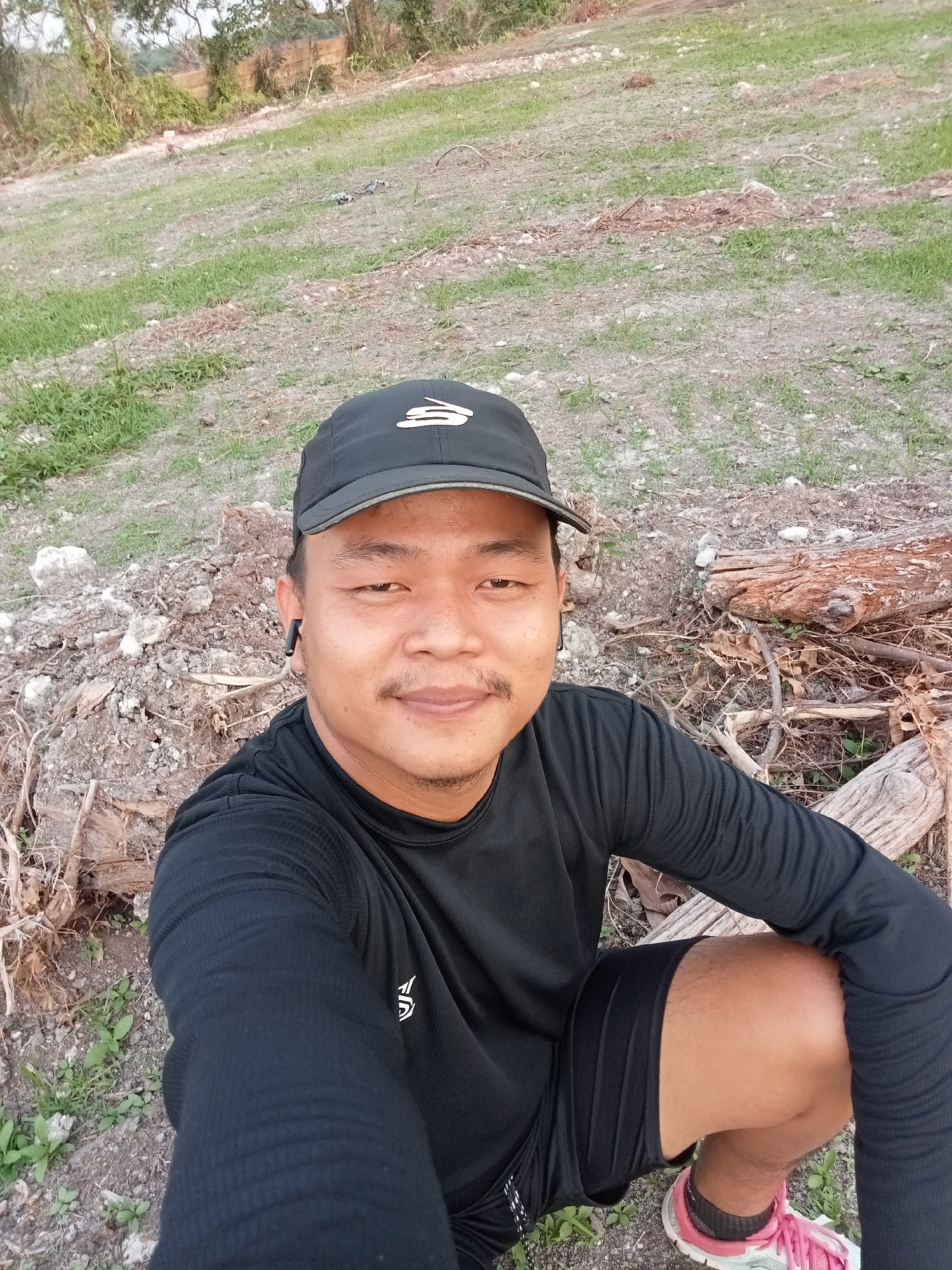 A young Asian man wearing a black long-sleeved shirt, black shorts, a black baseball cap with a white logo, and pink athletic shoes poses for a selfie outdoors. He is sitting on the ground in what appears to be a rural area. The background shows dry earth, patchy grass, and some fallen logs. The man has short dark hair, a mustache, and is wearing black earbuds. The lighting suggests it is daytime, likely late afternoon or early morning due to the soft light and long shadows. This location is consistent with the rural outskirts of Teluk Kenidai, Indonesia, characterized by open land and natural vegetation.
