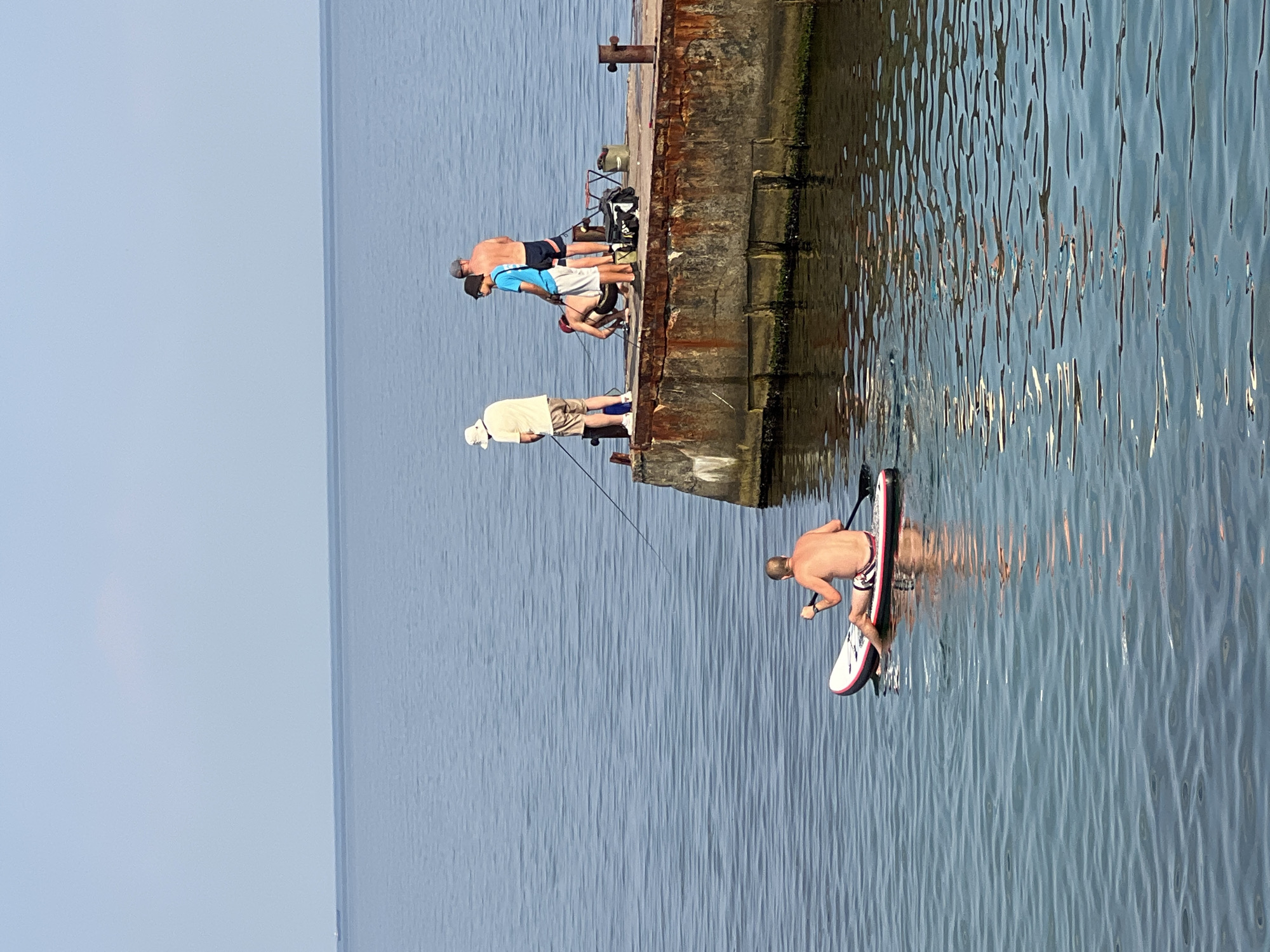The image depicts a bright, clear day at sea, likely in Odesa, Ukraine, given the context. In the foreground, a shirtless man with short hair and patterned shorts is seated on a stand-up paddleboard, facing away from the camera and holding a paddle. He is paddling in the calm, blue water, which reflects the light in rippling patterns.
In the mid-ground, a concrete pier juts out into the water. On the pier, four individuals are visible. Two men are standing, one in a yellow t-shirt and beige shorts, the other shirtless in blue shorts. Another man, also shirtless and wearing dark shorts, is crouched down, possibly fishing. A fourth person, wearing a blue t-shirt and white shorts, is standing and looking towards the other individuals. A fishing rod extends from near the standing man in the yellow shirt, and fishing gear, including a bucket and a bag, is present on the pier. The pier itself appears weathered and aged, with visible rust and concrete damage.
The background consists of a vast expanse of calm, blue water meeting a pale blue, cloudless sky. The horizon line is clear and sharp. The overall scene conveys a sense of leisure and outdoor activity on a warm day.