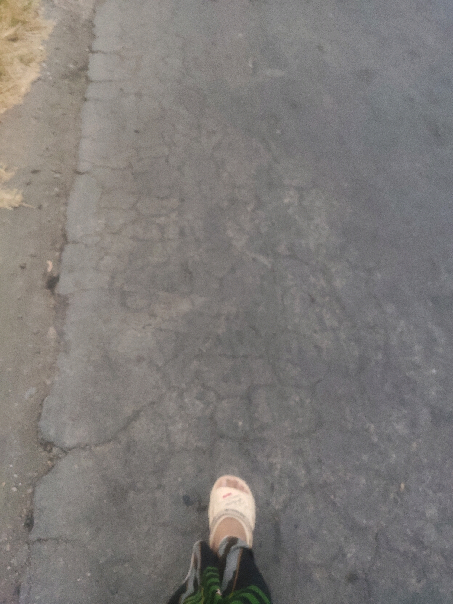 The image shows a person's feet wearing white sandals and dark-colored pants with green stripes. The person is standing on a cracked asphalt road. The road is gray and shows signs of wear and tear. To the left of the road, there is a patch of dry grass and some dirt. The scene appears to be outdoors. The location is described as Bongkot, Indonesia.