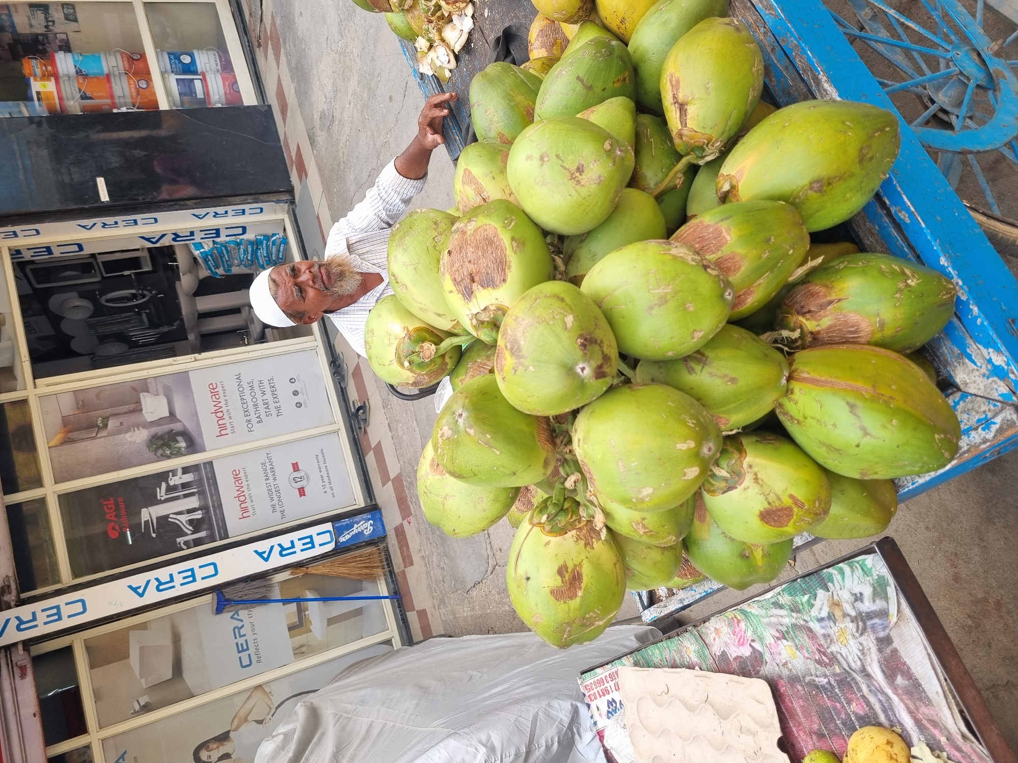 An adult male with a white beard and a white skullcap is seated next to a blue pushcart laden with green coconuts on a paved street in Puttaparthi, India. He wears a light-colored, striped shirt and has his right arm resting on the edge of the coconut cart. The cart holds a large quantity of whole green coconuts, many exhibiting brown blemishes, and a collection of husked coconut halves are visible on the far right. To the left of the coconuts, a table partially displays a patterned cloth and a few yellow fruits, alongside a white-covered object, possibly a vehicle. The background features a commercial storefront with large glass windows. Advertisements for "CERA," "AGL Bathware" (displaying faucets), and "hindware" (mentioning a "12 year warranty on all Hindware faucets") are visible on the windows, which also showcase various white bathroom fixtures. Further right within the shop's interior, stacks of colorful paint buckets, including the "NEROLAC" brand, are visible. A blue plastic mop and a straw broom lean against a vertical section of the storefront. A pair of dark sandals are on the pavement near the shop's entrance.