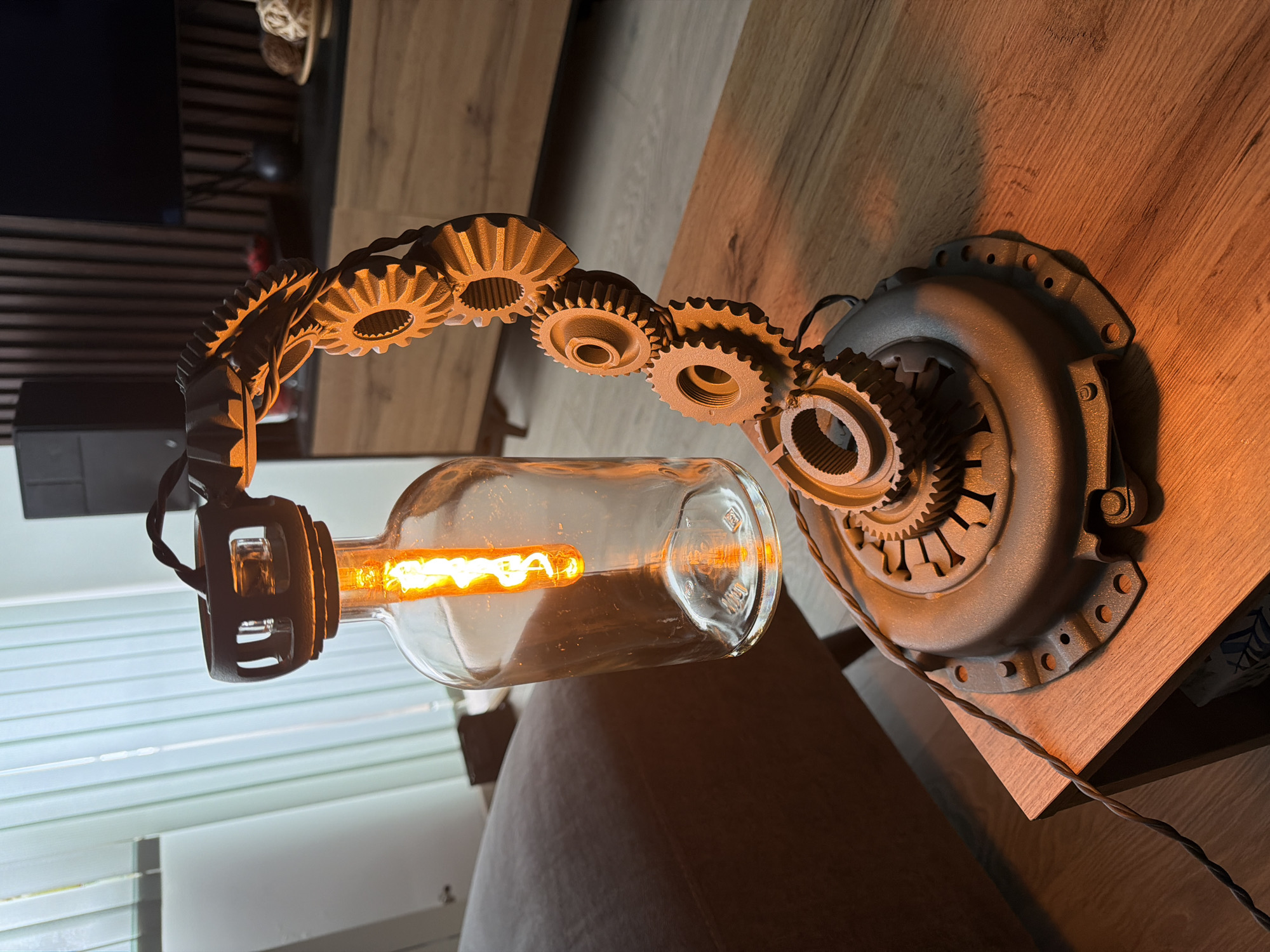 A creatively designed steampunk-style table lamp is prominently displayed on a light-colored wooden surface, likely a table or desk. The lamp's base appears to be a repurposed metallic grey automobile clutch cover or pressure plate, featuring several bolt holes around its circumference. Extending from the base is an articulated arm fashioned from a series of interconnected gears of varying sizes, also painted a consistent metallic grey, some with a subtle bronze highlight from the lamp's glow.
At the end of the gear-assembly arm, a clear glass bottle, reminiscent of a liquor or beverage bottle, cradles an Edison-style light bulb. The bulb emits a warm, bright orange-yellow light, showcasing its intricate filament. A black electrical cord with a twisted fabric sheath runs from the lamp's base along the wooden surface.
The background reveals an indoor setting. A dark rectangular object, possibly a television or monitor, and horizontal wooden slats, likely part of furniture or wall paneling, are visible behind the lamp. To the left, white horizontal window blinds allow some diffused light into the room. The overall lighting suggests an evening or indoor environment where the lamp provides primary illumination.
Visible text on the glass bottle includes "200ML" and some other faint, unreadable embossed characters. No people are present, and the scene focuses entirely on the unique, industrial-themed lamp.
