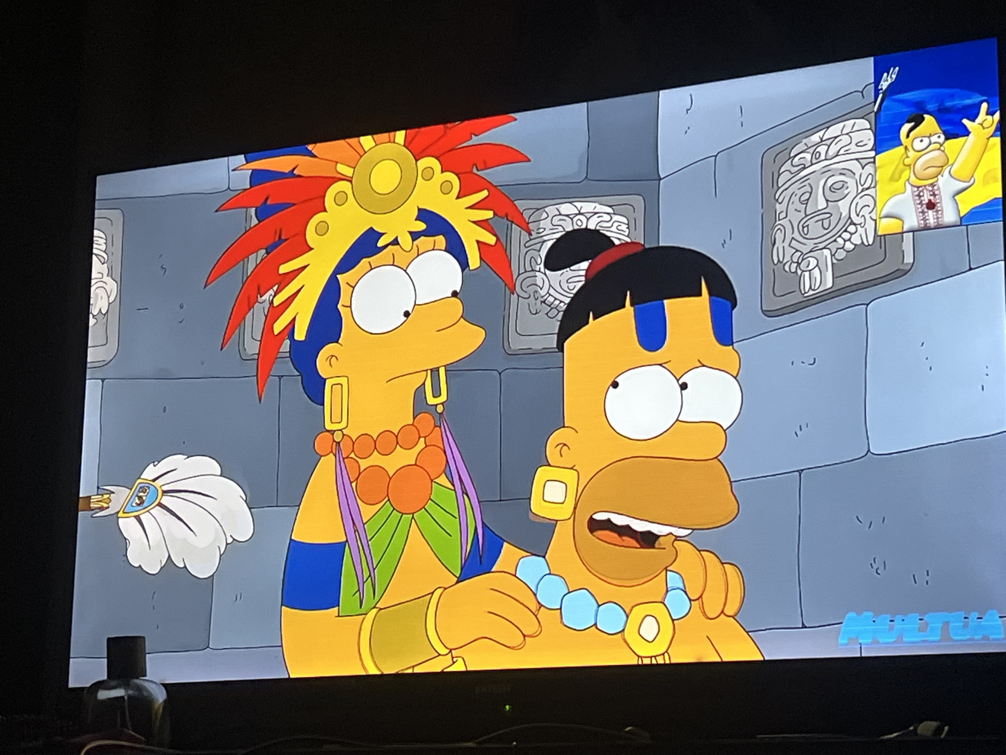 The image is a screenshot from a television screen showing an episode of The Simpsons. The characters Lisa Simpson and Homer Simpson are depicted in what appears to be an ancient Mayan or Aztec setting, wearing traditional headdresses and jewelry. The background consists of stone walls with carvings. A small image of Homer Simpson in a Ukrainian embroidered shirt, giving a peace sign with the Ukrainian flag behind him, is superimposed in the upper right corner. The scene is likely from a fictional depiction within the show and has no connection to the city of Zoriane, Ukraine.