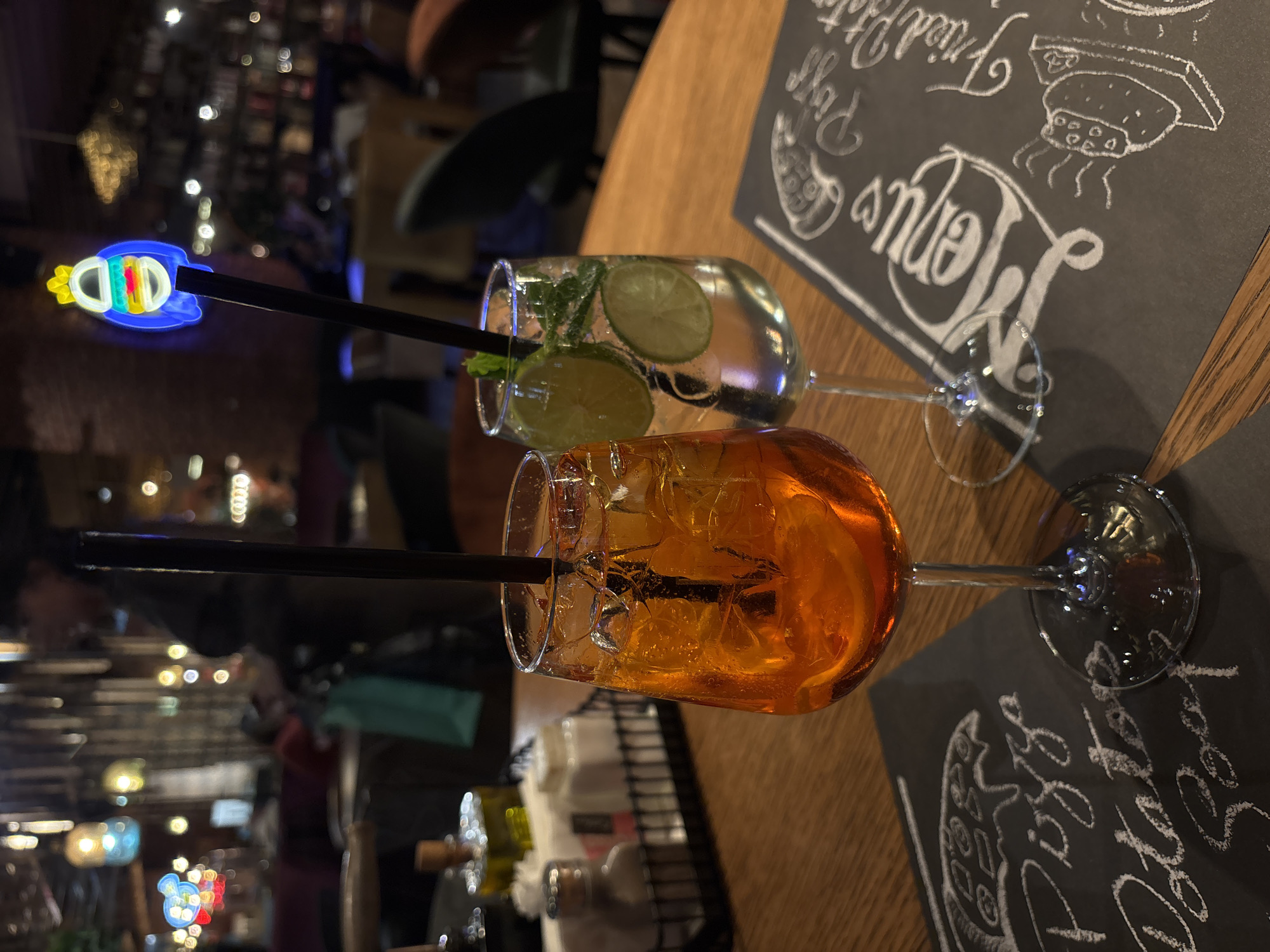 The image captures a dimly lit interior of what appears to be a restaurant or bar. A wooden table takes prominence, hosting two stemmed cocktail glasses. One holds a clear liquid with slices of lime and mint leaves, while the other contains an orange-colored drink with ice. Both are equipped with black straws, with a neon light-up adornment on the lime drink straw. Below the drinks, condiments sit on the table. The background features blurred lights and shapes, suggesting a busy environment. The table is covered with dark paper containing the words “Venus”, “Pizza”, and “Fried Potatoes” written on it in white chalk. Based on this, it is likely that this photo was taken at the Venus Restaurant in the city of Varna, Bulgaria.
