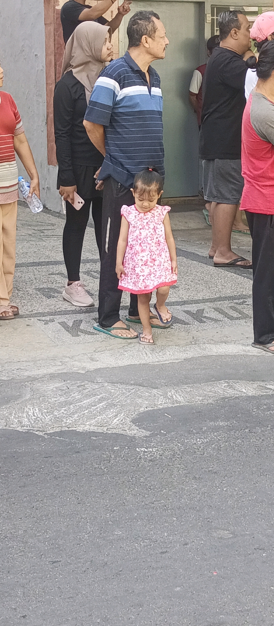 A group of people are standing on a patterned concrete surface outdoors in what appears to be a city street or public area in Malang, Indonesia. The group includes a young girl in a pink floral dress with her hair tied back in a ponytail. She is wearing flip-flops and looking down. To her left, an adult male in a blue and white striped polo shirt and dark pants is looking to the right. Behind him, a woman in a light brown hijab and black clothing is also looking to the right. To the right of the girl, more people are visible, including a person in a red shirt and another in dark shorts. A water bottle is held by a person on the far left of the image. The lighting suggests it is daytime.
