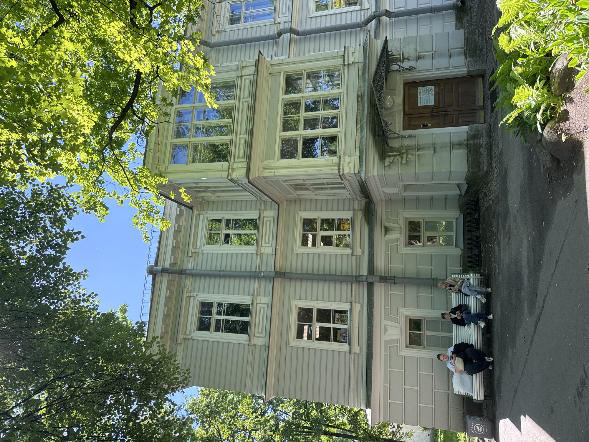The image depicts the exterior of a light-colored, multi-story building, likely a historical structure, located in Saint Petersburg, Russia. The building features numerous windows with white frames and is partially obscured by lush green trees casting shadows. In front of the building, three individuals are seated on a white bench, each engaged with their mobile phones. To the left of the bench, a grey trash receptacle with a circular logo is visible. The ground in front of the building is paved, with a patch of green ferns and rocks in the foreground. The overall scene suggests a quiet, daytime setting in a park or historical district.