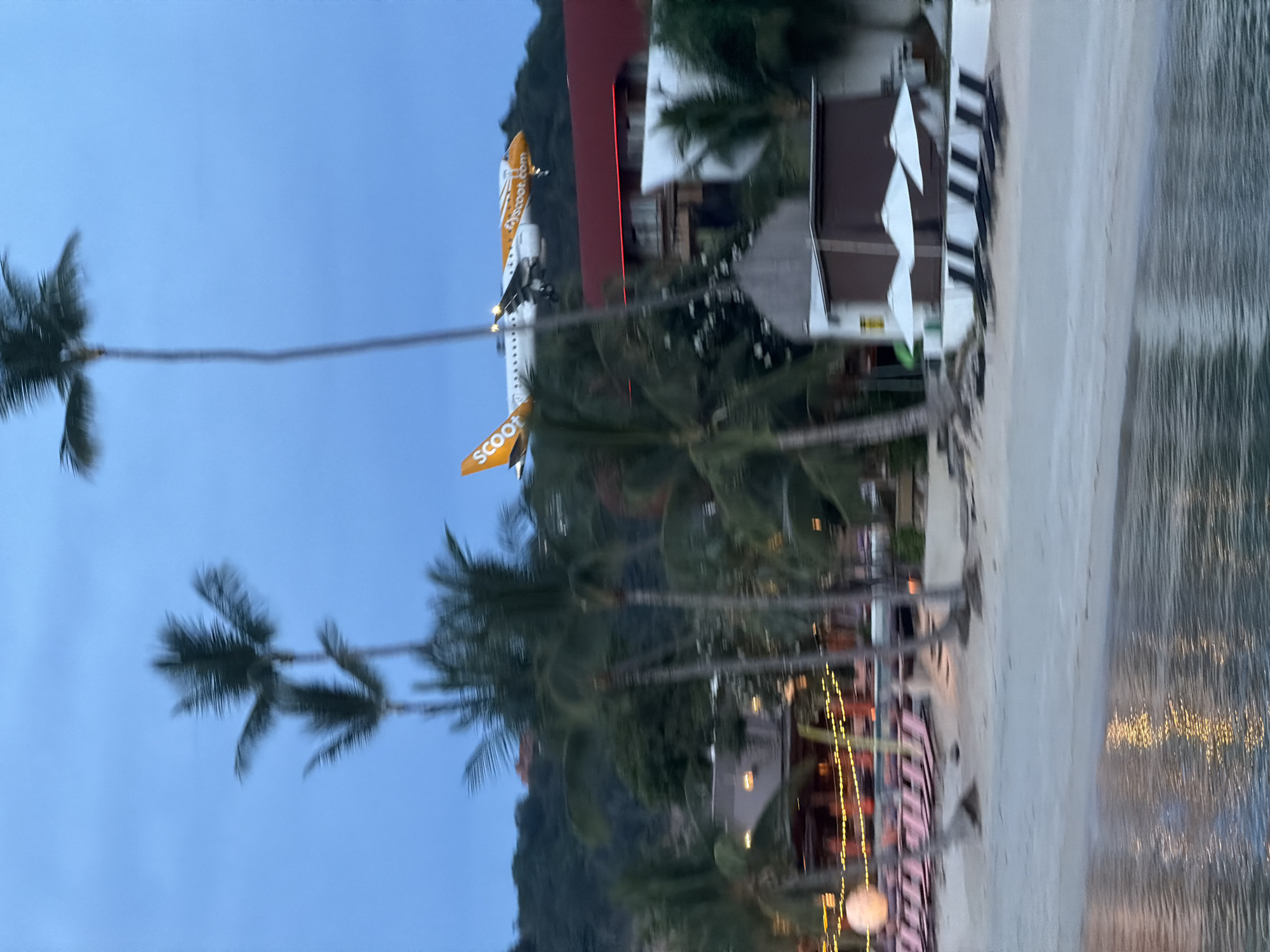 The image depicts a beach scene at dusk or early morning in Baan Bang Rak, Thailand. In the sky, an orange and white airplane with the word "SCOOT" visible on its tail is descending, likely on approach to an airport. The plane is positioned above a lush, green hillside covered with trees and some buildings. In the foreground, there is a beach with rows of empty lounge chairs and umbrellas. Palm trees are scattered throughout the scene, some with their fronds blurred due to movement or the image quality. The water in the foreground reflects the lights from the beach area, creating a shimmering effect. The overall atmosphere is tranquil, with a hint of activity from the approaching aircraft.
