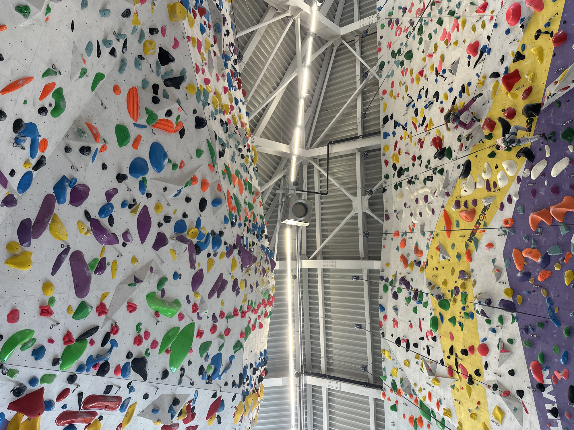 The image shows an indoor climbing gym in an unknown location. Two climbing walls with numerous colored climbing holds are visible, reaching up to a ceiling of metal girders and bright overhead lights. No people are visible in the shot.